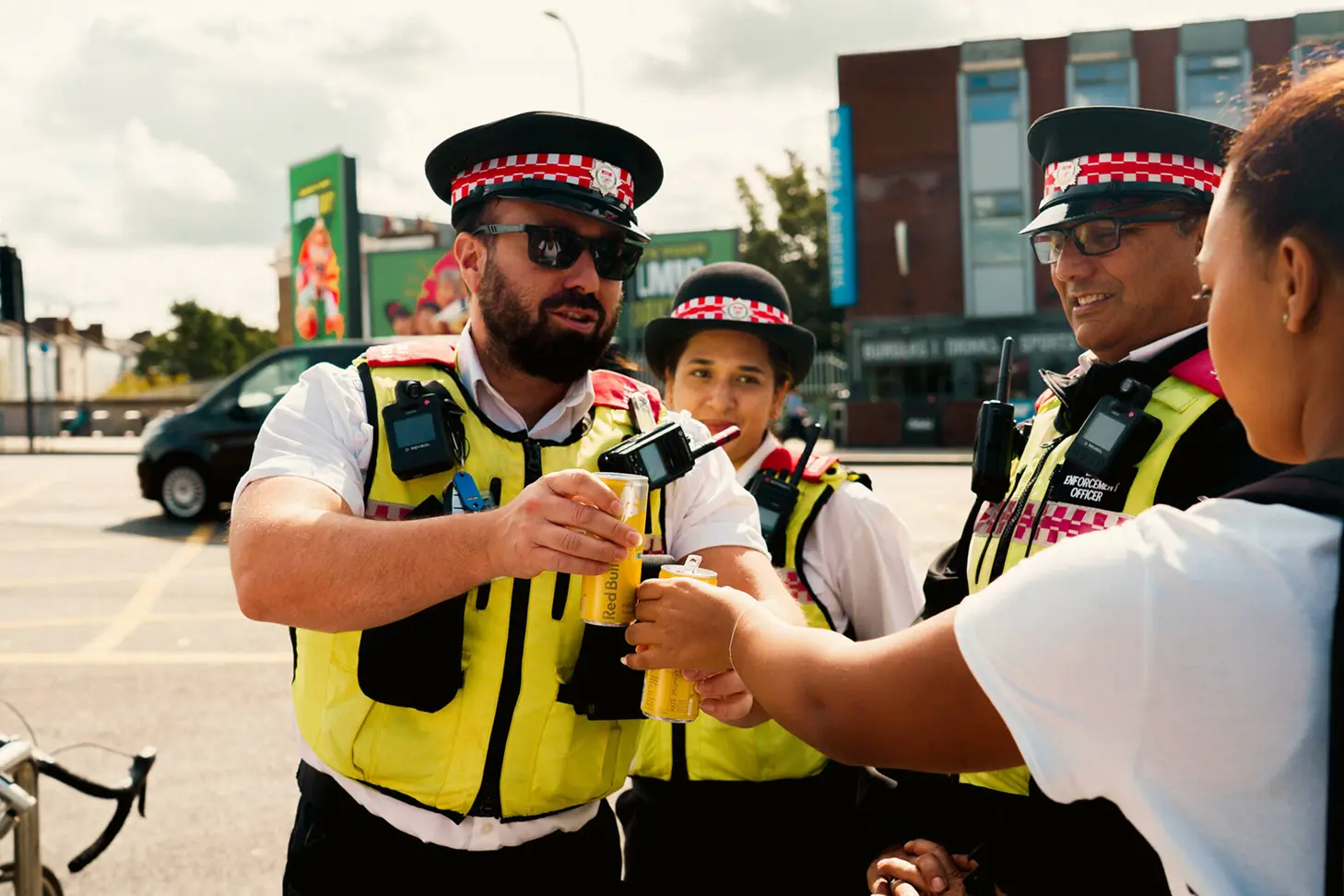 Three uniformed enforcement officers wearing high-visibility yellow vests and black hats with red checkered bands are outdoors, clinking cans of Red Bull energy drink with a person in a white and black shirt.