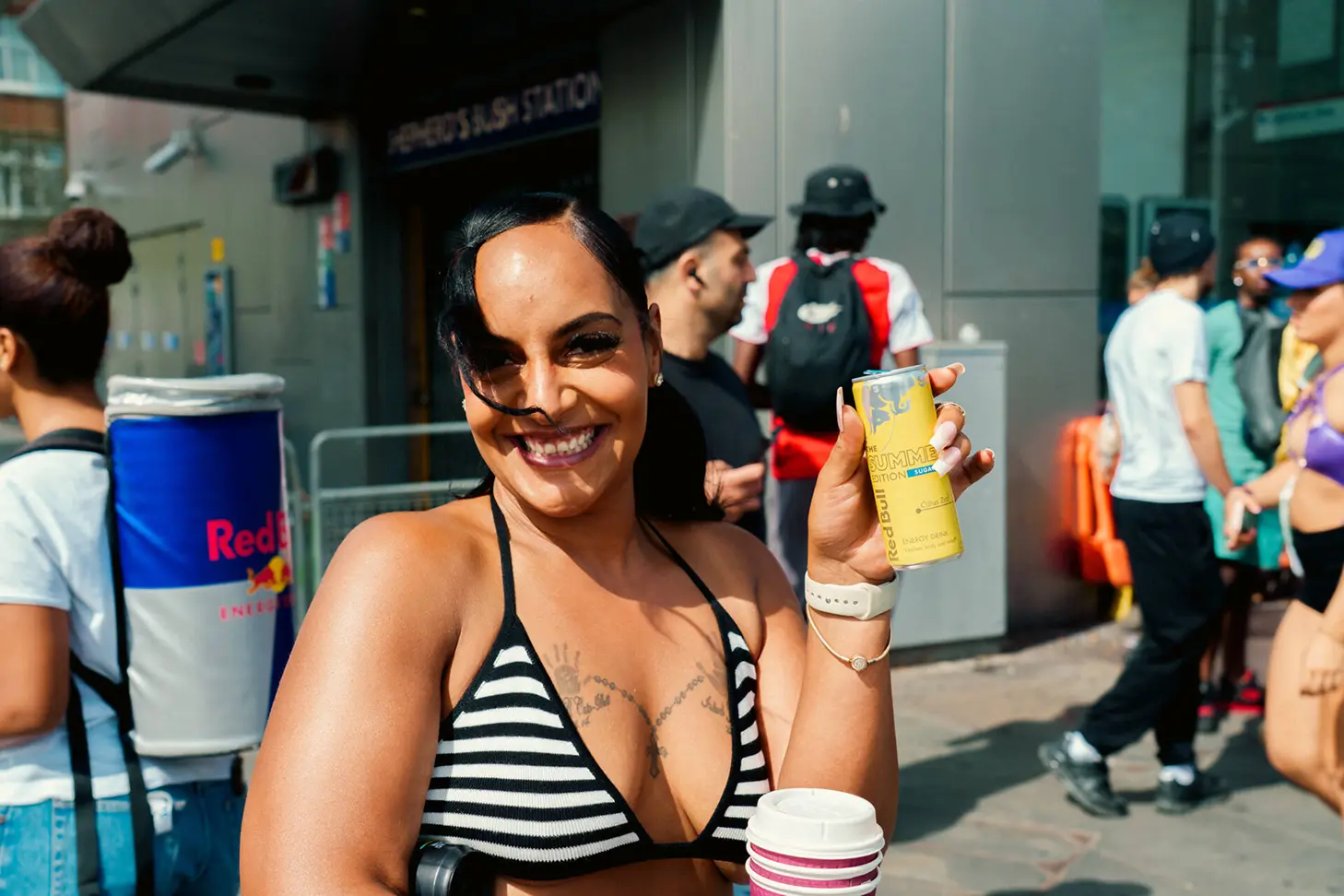 Smiling woman in a black and white striped bikini top holding a yellow can of Red Bull Summer Edition energy drink outdoors among people.