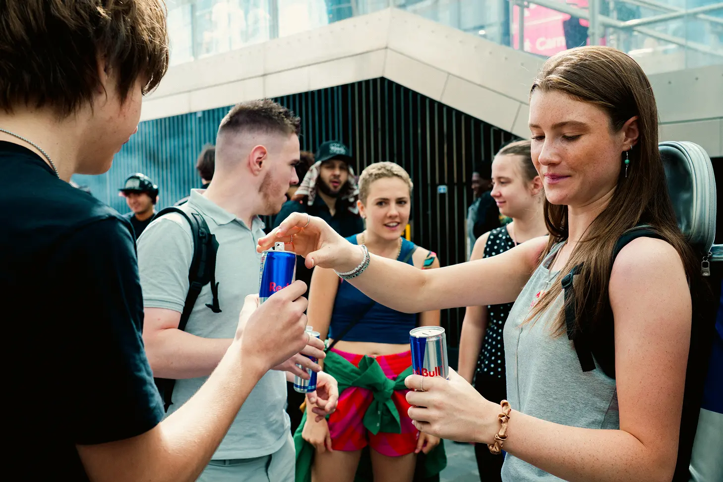 A group of young adults outdoors casually socializing while holding cans of Red Bull energy drink, with a modern urban background.