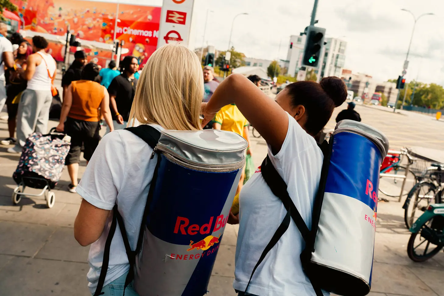 Two people wearing white shirts carrying large red and blue Red Bull energy drink backpacks walking on a busy city sidewalk with other pedestrians and bicycles around.
