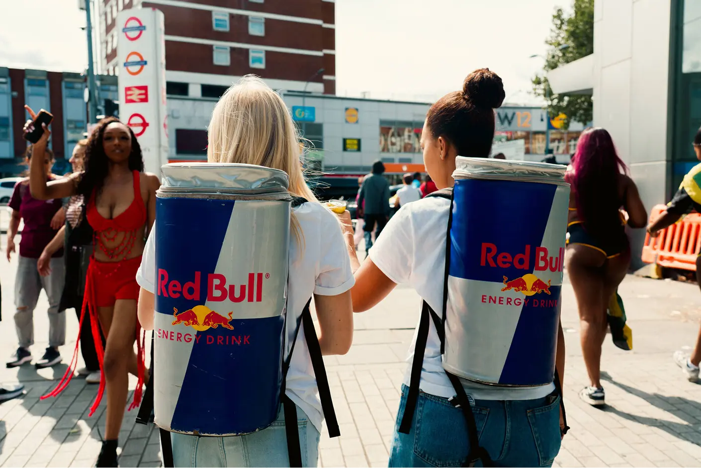 Two women wearing white T-shirts and jeans walking down a city street with large Red Bull energy drink can backpacks on their backs, surrounded by other pedestrians.