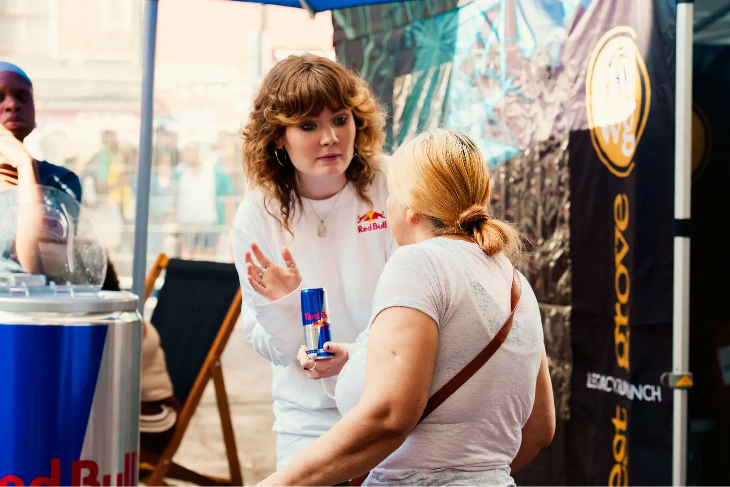 A woman in a white Red Bull shirt holding a can of Red Bull and talking to another woman with blonde hair tied back, inside a tent at an outdoor event with a Red Bull branded display and promotional materials visible.