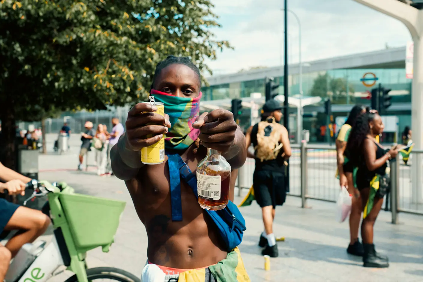 Shirtless man wearing a colorful face covering holds out a can and a bottle toward the camera on a city sidewalk with other people and a green bike in the background.