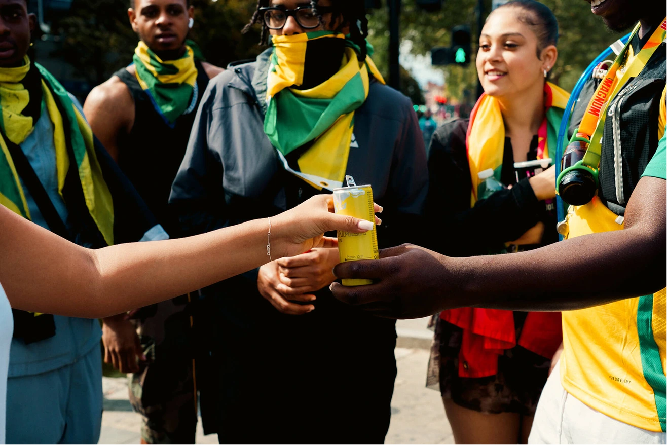 People wearing Jamaican flag-themed scarves and clothing at an outdoor event share a yellow can, with one person handing it to another.