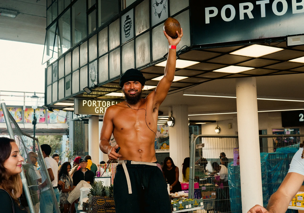 A shirtless man wearing a black head wrap and black pants is holding a coconut up with one hand while jump roping in a busy market area under a sign that reads PORTOBEL. People and market stalls are visible in the background.