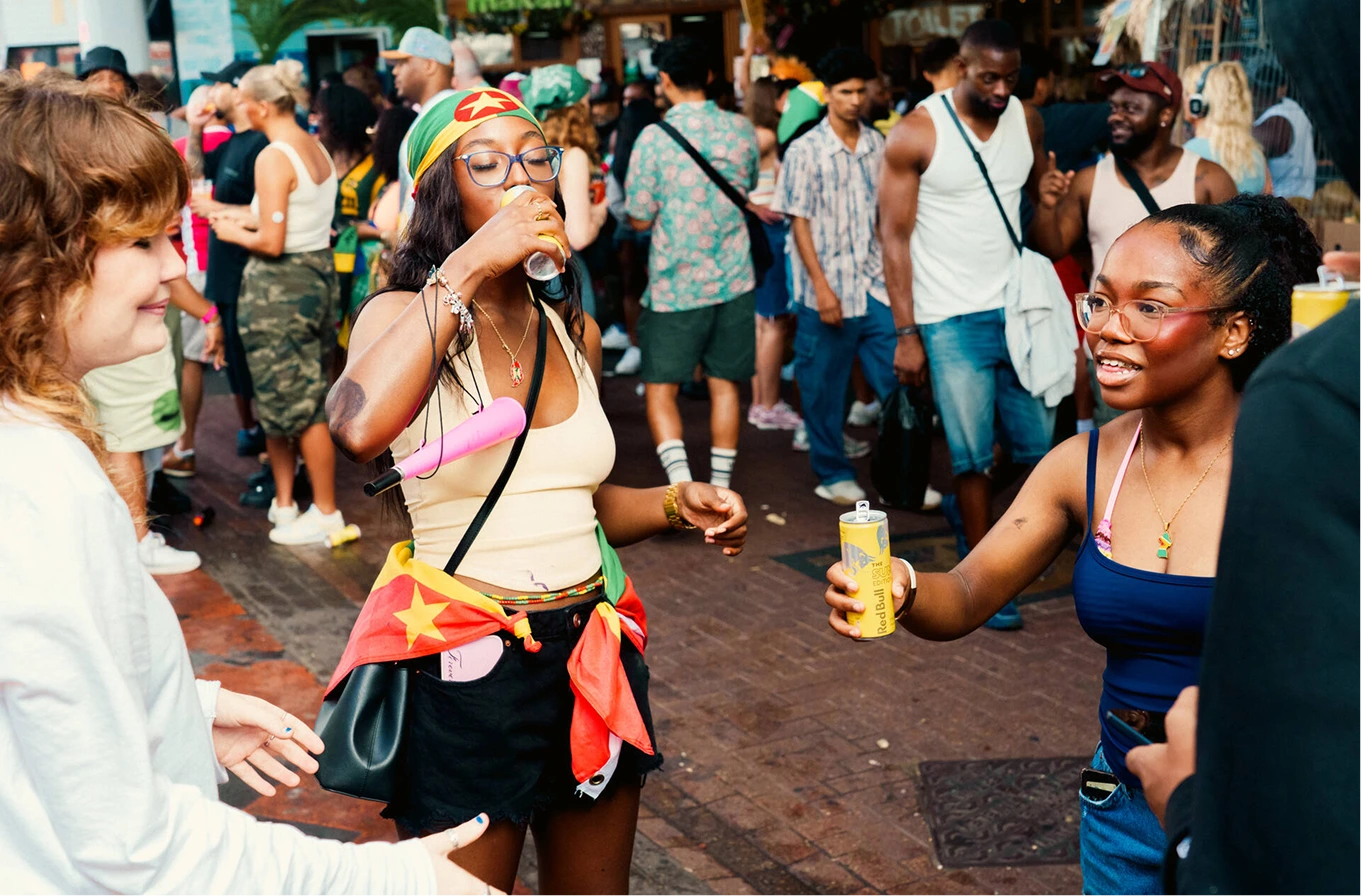 A lively street scene with three young women in the foreground enjoying drinks, one wearing glasses and a colorful headscarf with a star design, one in a blue top holding a can, and the third smiling with curly hair. The background shows a crowd of casually dressed people at an outdoor event with tropical plants visible.