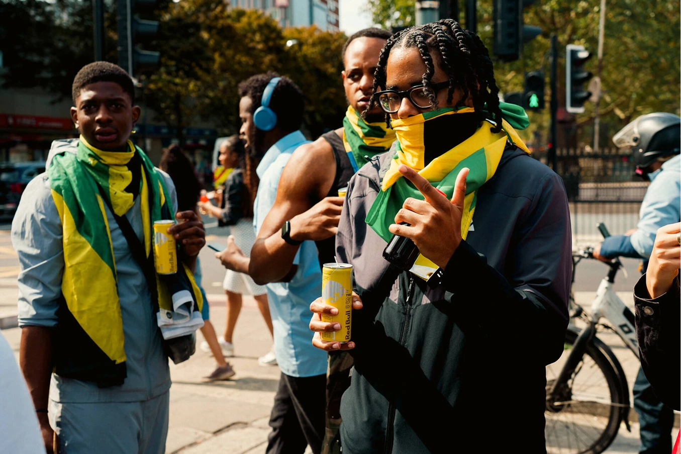 A group of young men outside on a sunny day, some wearing yellow-green-black scarves resembling the Jamaican flag and holding yellow cans of Red Bull energy drink, with a person in the background wearing blue headphones and another riding a bicycle.