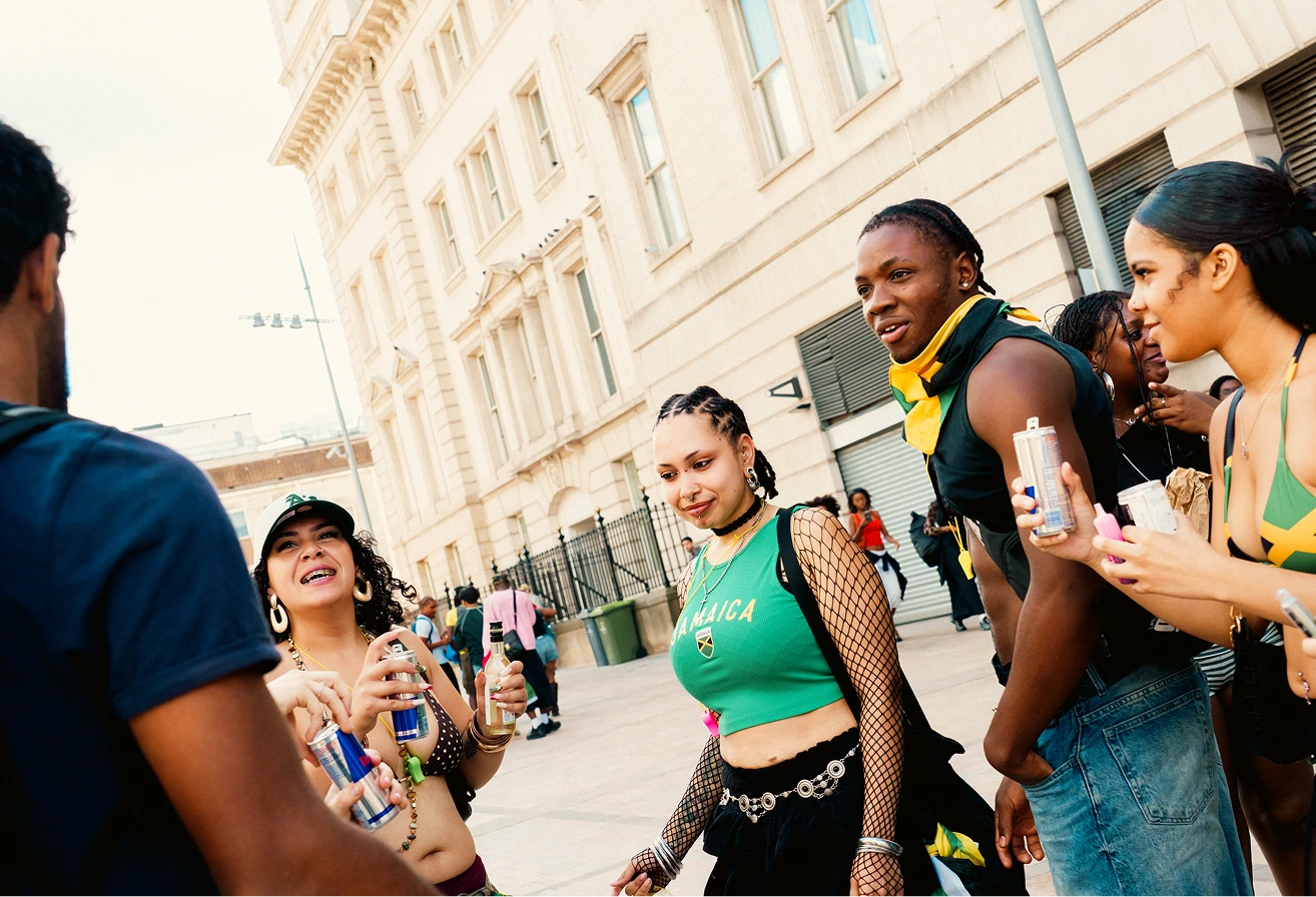 A group of young people outdoors on a city street in front of a large building, socializing and holding drinks with some wearing clothing featuring the Jamaican flag colors and symbols.