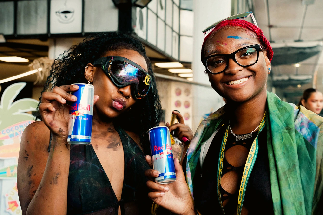 Two young women smiling and holding cans of Red Bull energy drink in an indoor setting; one woman has curly hair, wears reflective goggles, and the other has red braided hair, glasses, and colorful paint on her face.