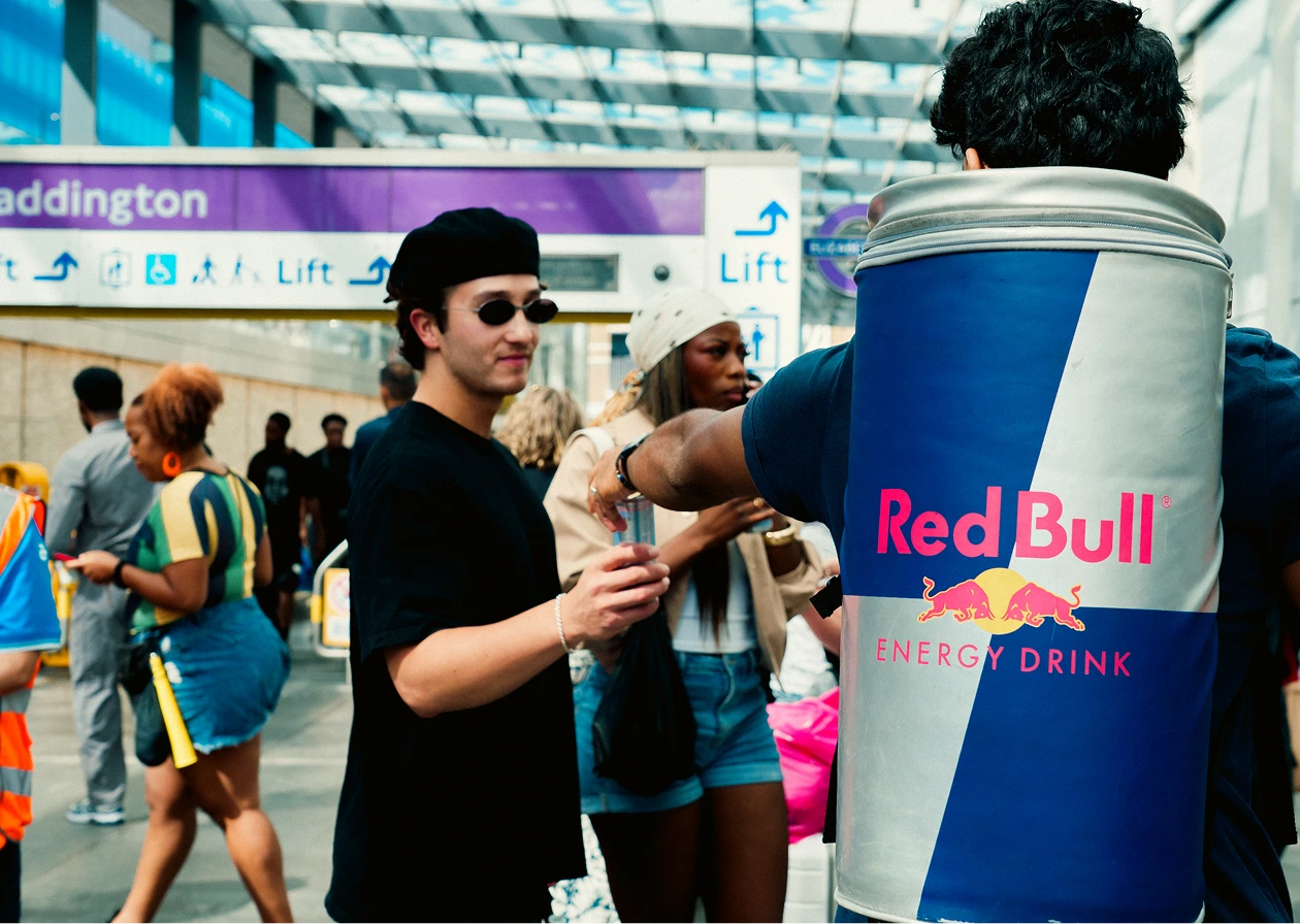 A person dressed as a giant Red Bull energy drink can hands a can to a man in black wearing sunglasses and a black beret at a busy indoor transportation hub with a Paddington sign in the background.