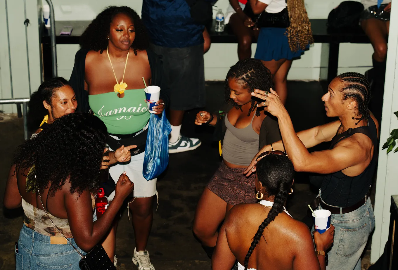 A group of six people dancing and socializing indoors at a party, holding drinks and wearing casual clothes, with one woman wearing a green 'Jamaica' top and a man with cornrow braids animatedly gesturing.