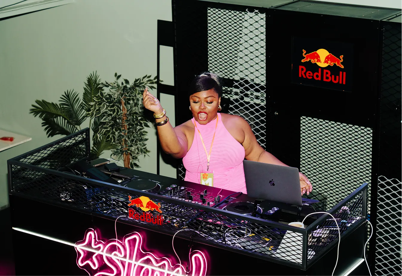 Female DJ wearing a pink sleeveless top performing energetically behind a DJ booth decorated with Red Bull logos and pink neon lights.