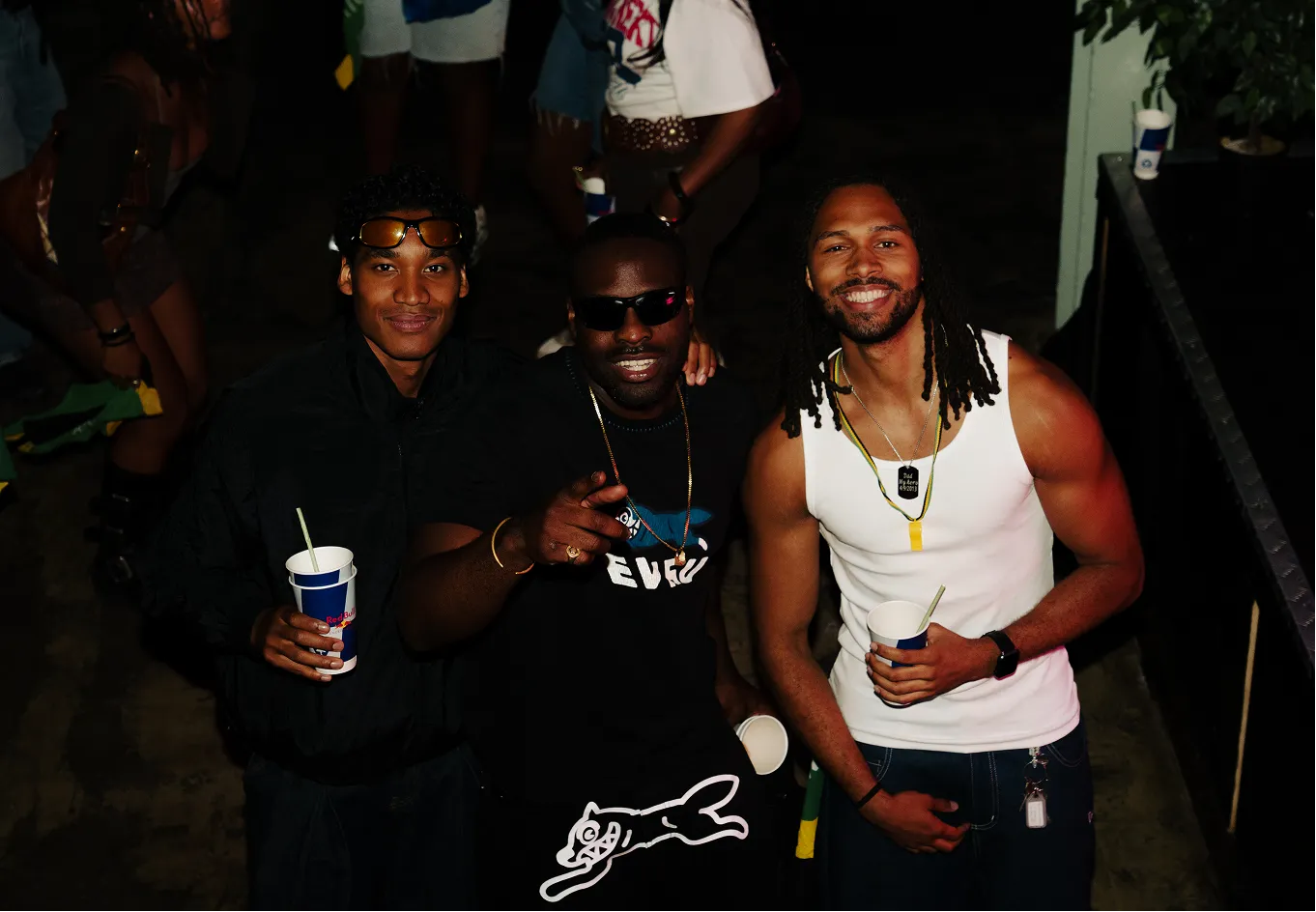 Three men posing and smiling at a night event, each holding a Red Bull cup; the man on the left wears sunglasses on his head and a black jacket, the man in the middle wears dark sunglasses and a necklace, pointing at the camera, and the man on the right with dreadlocks wears a white tank top and an Apple Watch.