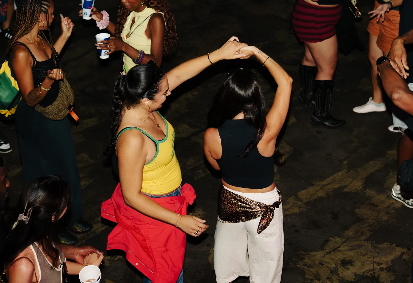 Two women dancing together in a crowd at a party or event, one wearing a yellow top with a red jacket tied around her waist and the other in a black top with white pants and a leopard-print scarf tied around her waist.