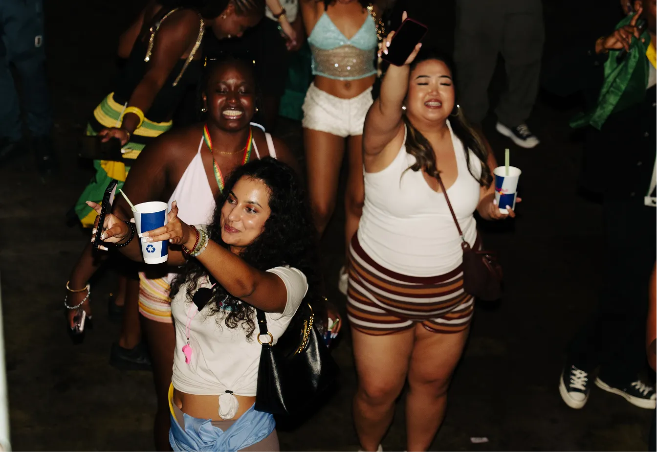 Group of diverse young women dancing and taking selfies at a nighttime event, holding drinks in blue and white cups.
