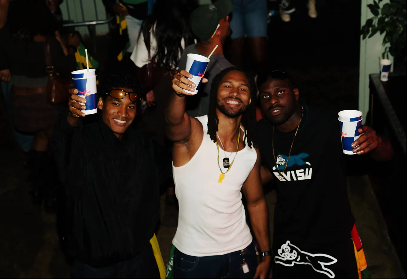 Three smiling men holding up Red Bull cups in a celebratory toast at a social event.