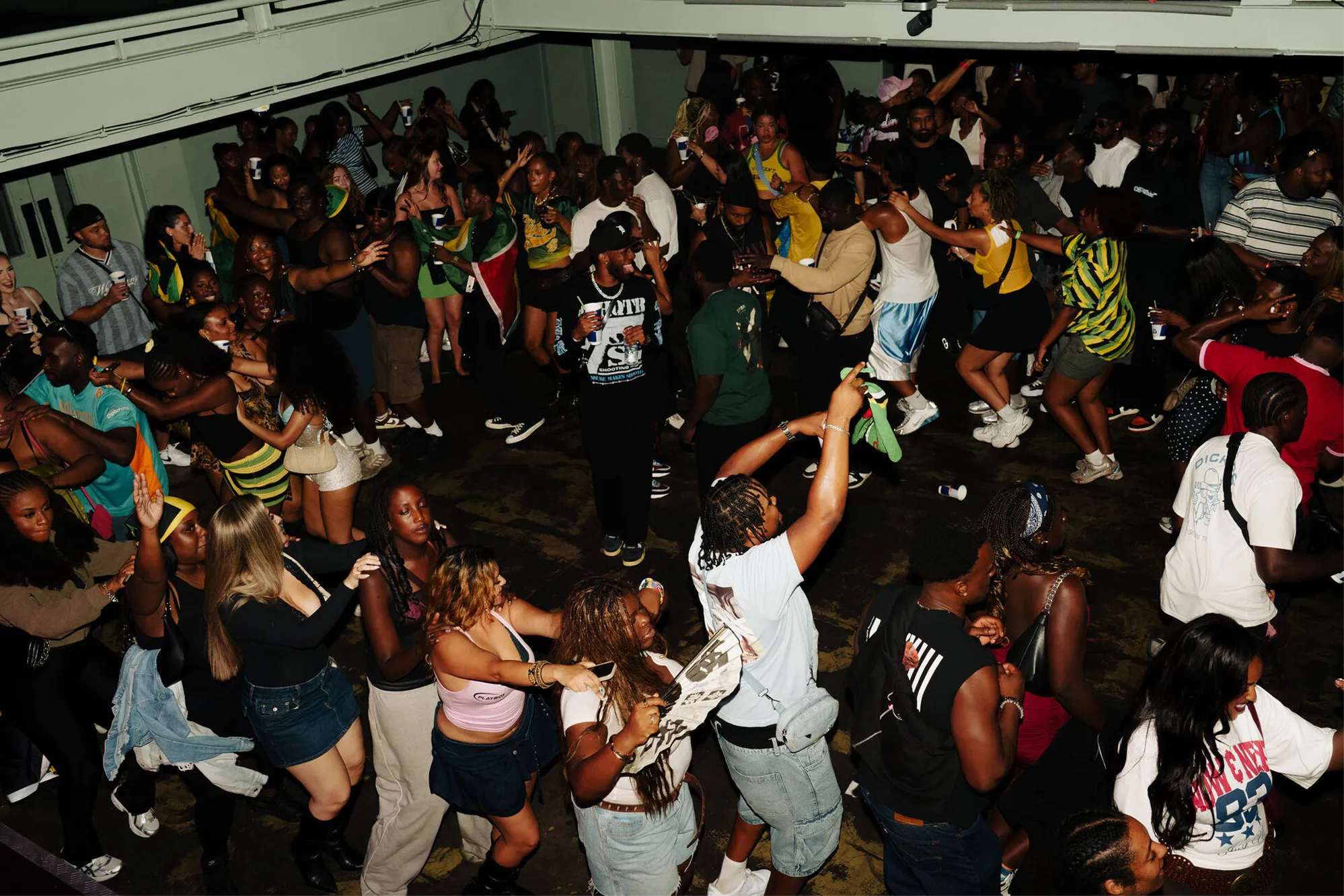 Crowded indoor party scene with diverse people dancing and socializing closely together, some holding drinks, under a low ceiling.