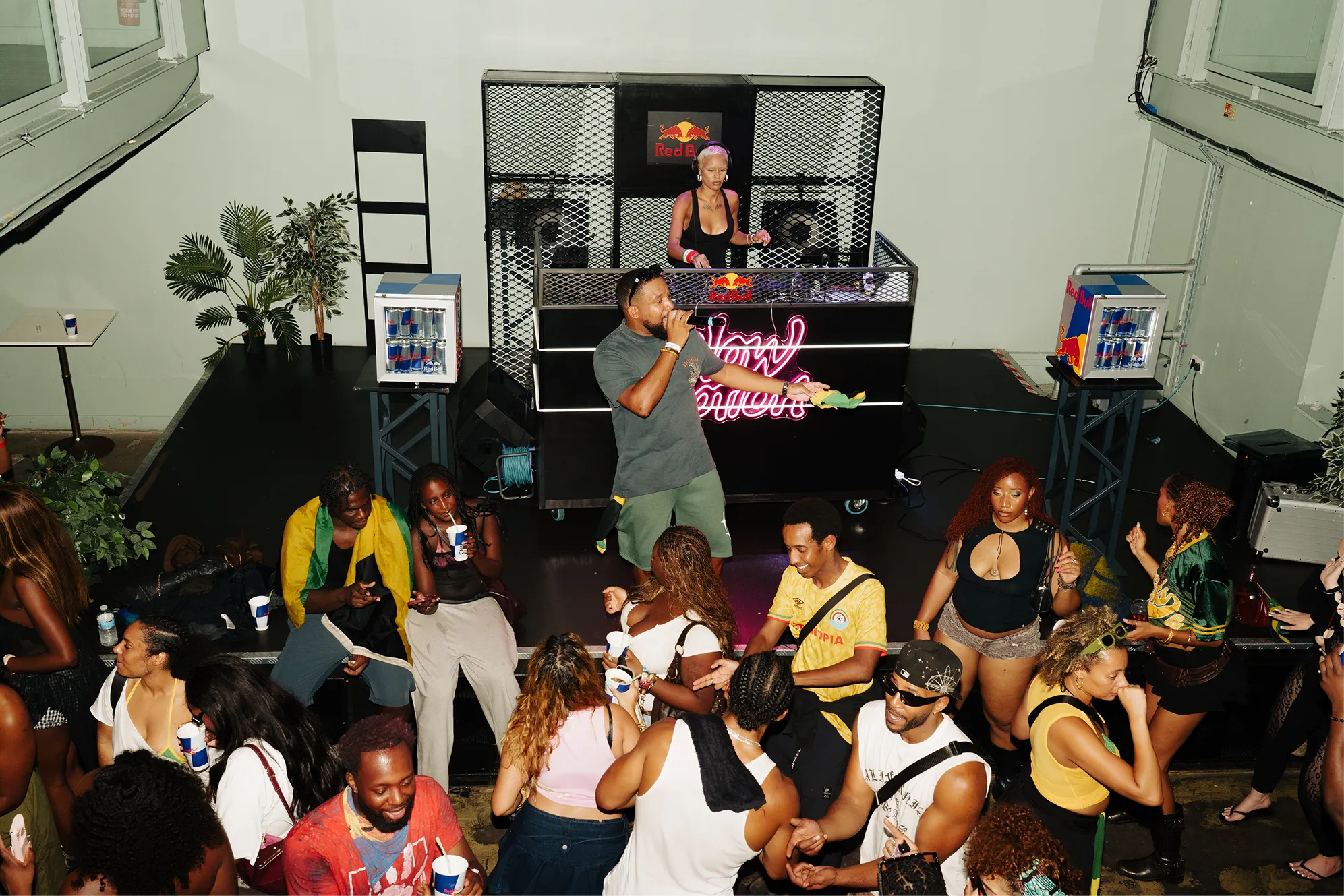 Crowd of people enjoying a lively music event where a male singer performs in front of a DJ booth with a female DJ mixing music, flanked by Red Bull mini-fridges on both sides.