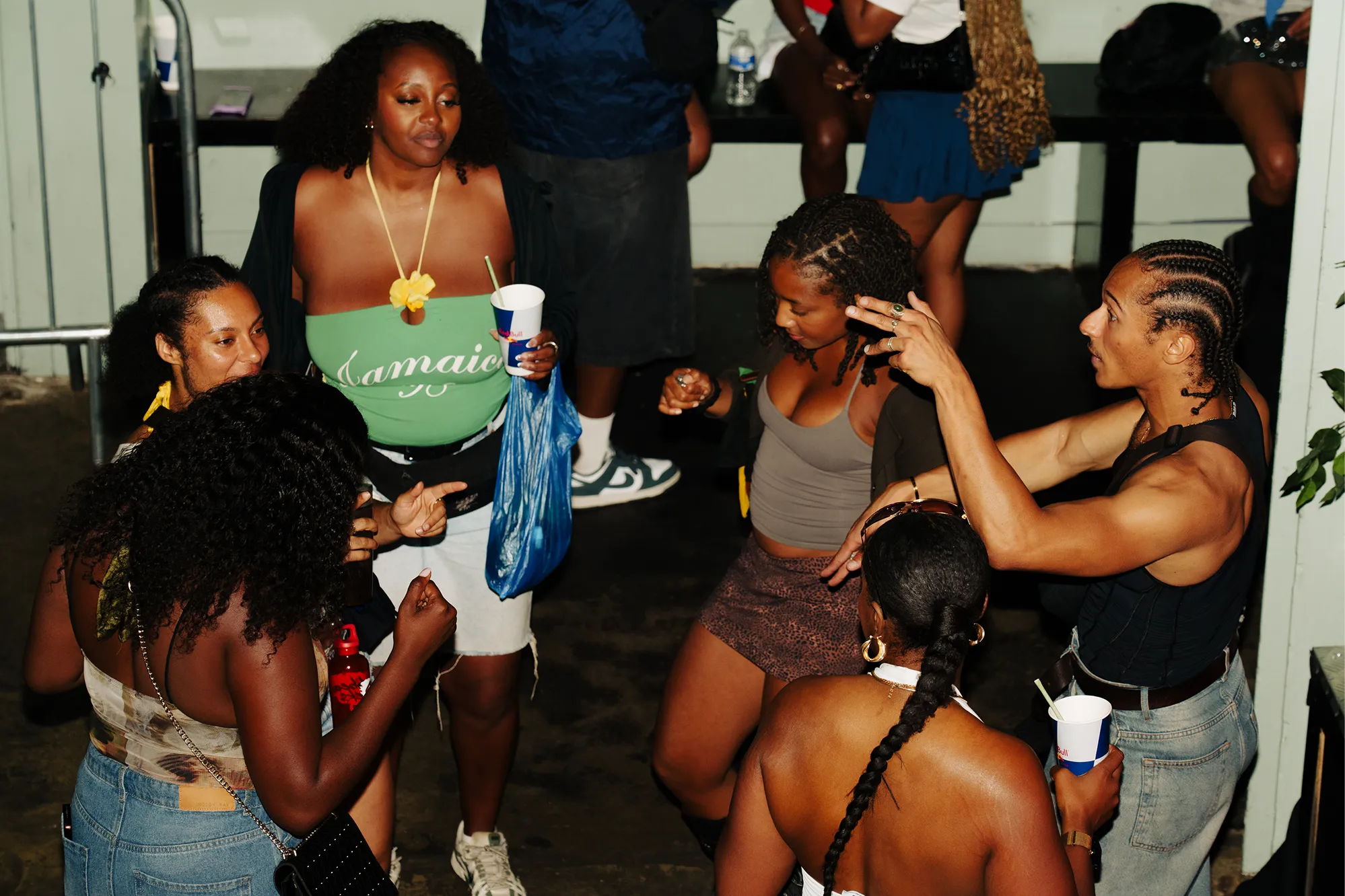 A group of six diverse people socializing and dancing indoors, some holding cups, with the person in the center wearing a green top with 'Jamaica' written on it.