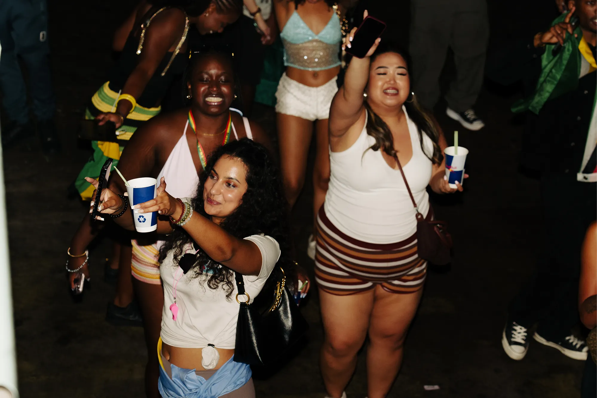 A group of young women at a nighttime event are raising their smartphones and holding drinks in cups with straws, smiling and appearing to enjoy the moment.