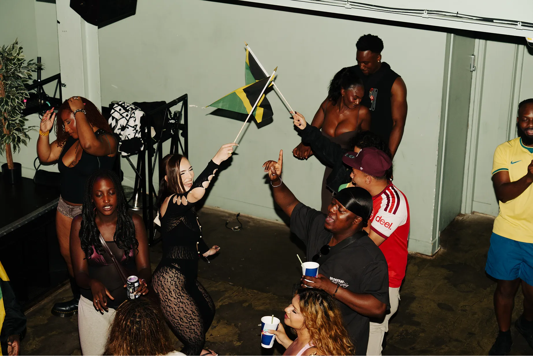 A lively group of people dancing and enjoying a party indoors, where two women are waving small Jamaican flags while others hold drinks and smile.