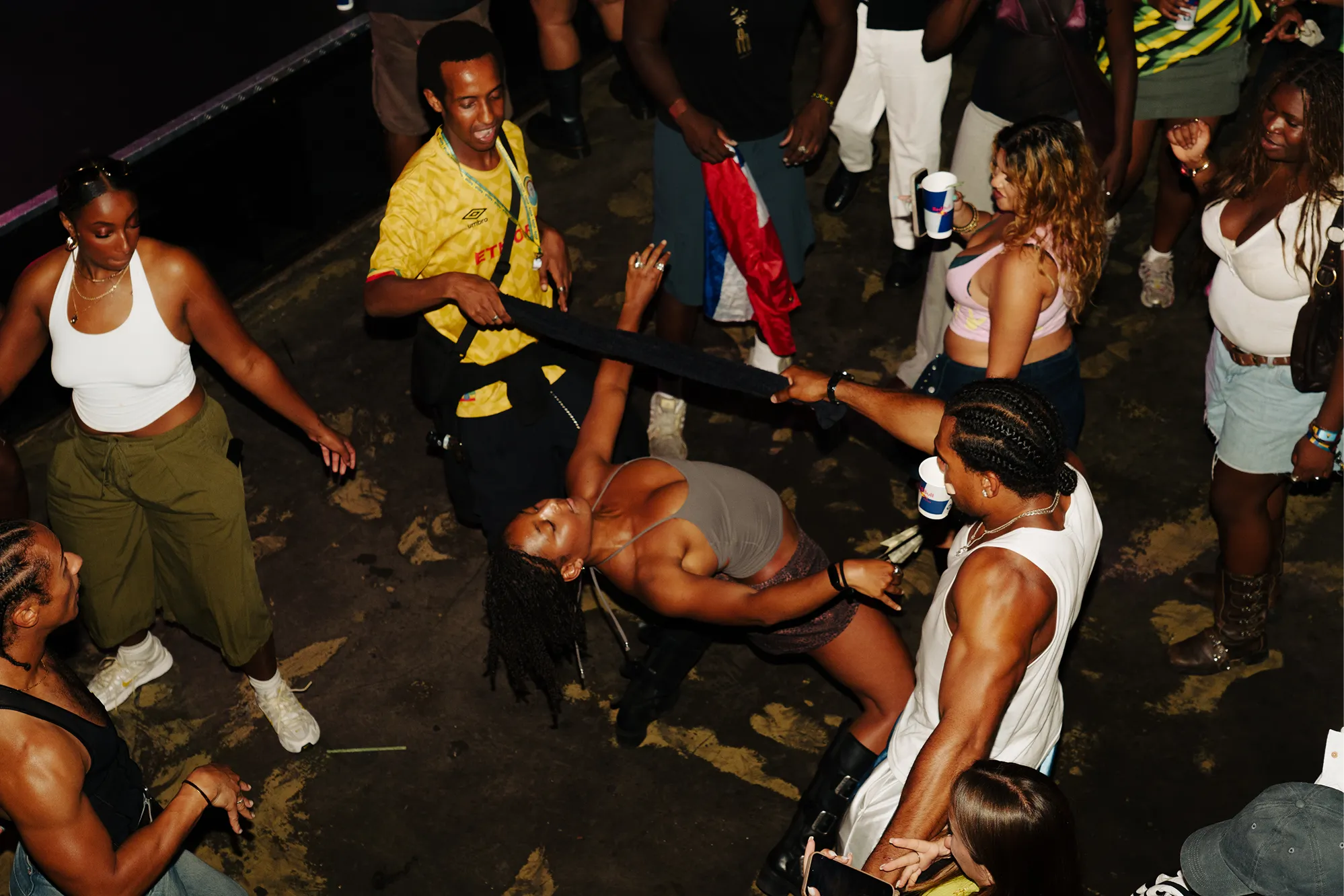 Group of people dancing indoors, with one woman bending backward under a held black fabric, surrounded by others watching and moving, some holding drinks.