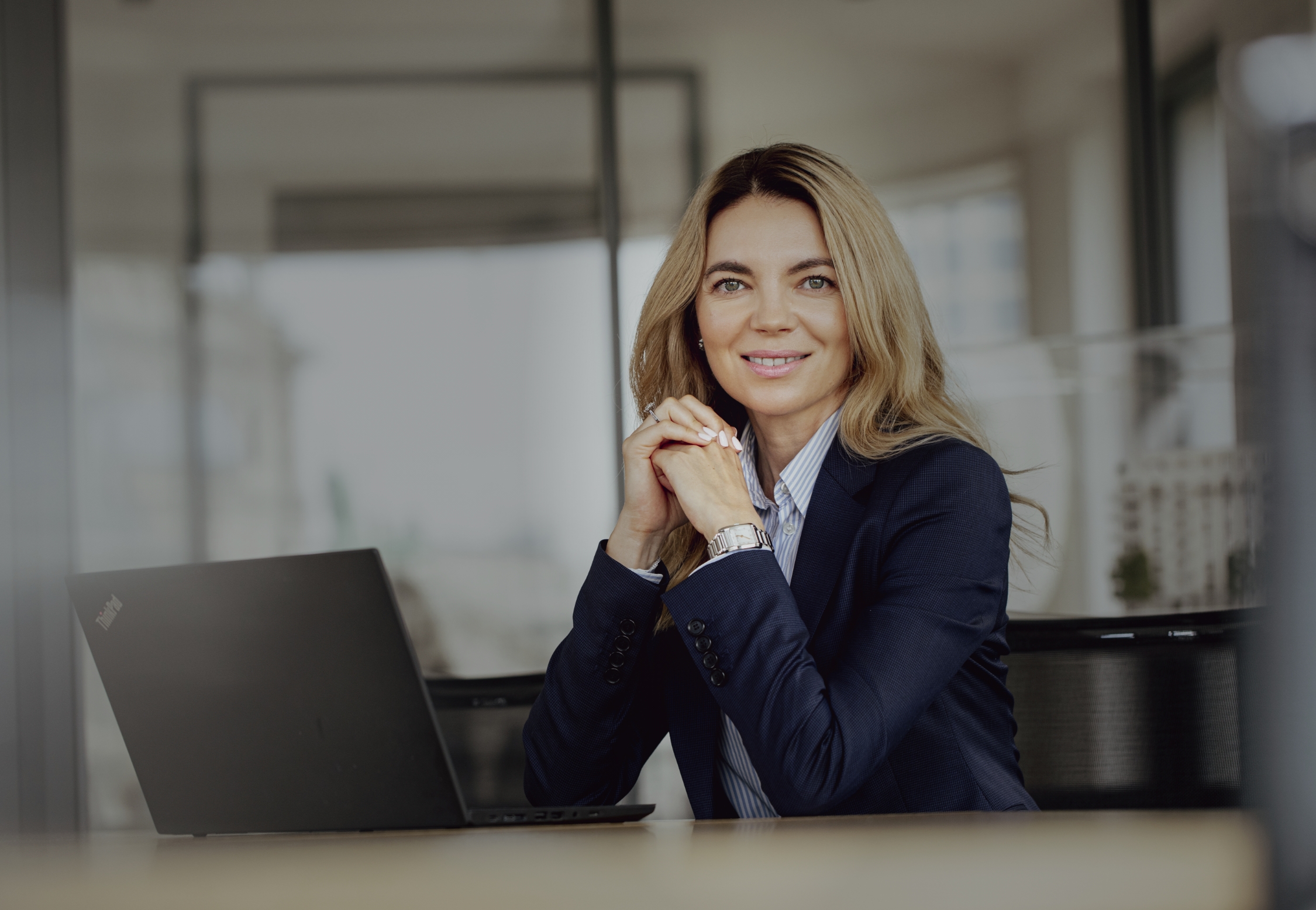 Smiling woman in a navy blazer sitting at a desk with a laptop in a modern office.