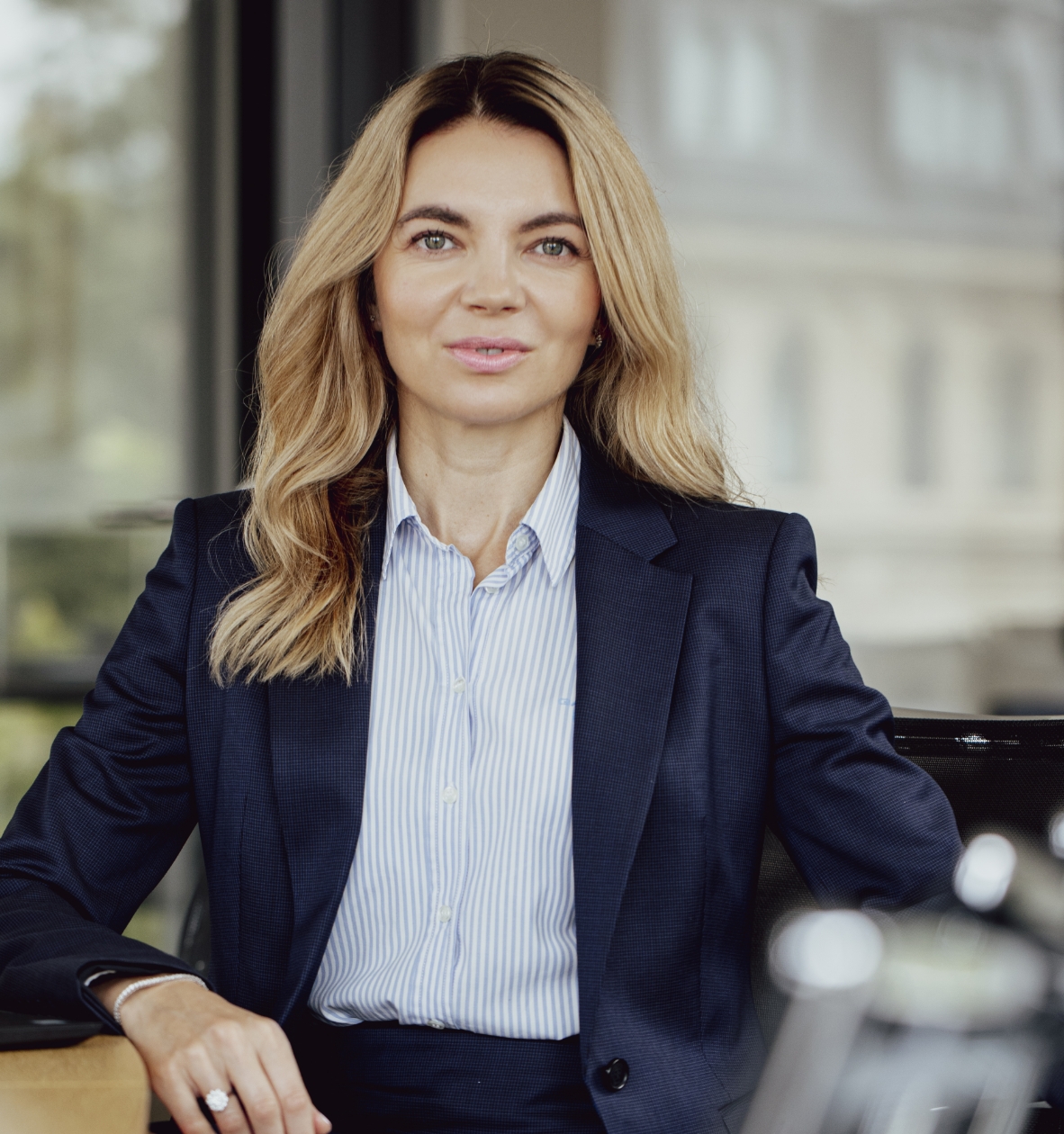 Professional woman with long blonde hair wearing a navy blazer and striped shirt sitting indoors.