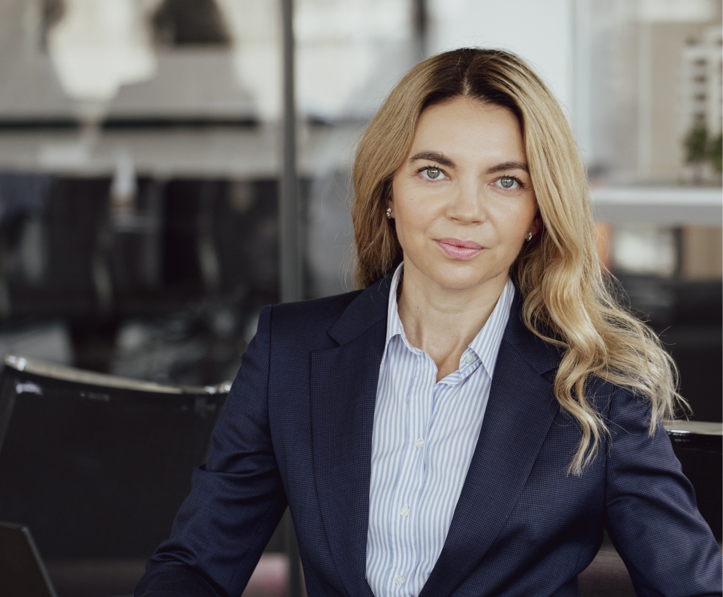 Professional woman with long blonde hair wearing a navy blazer and striped shirt, sitting in an office.