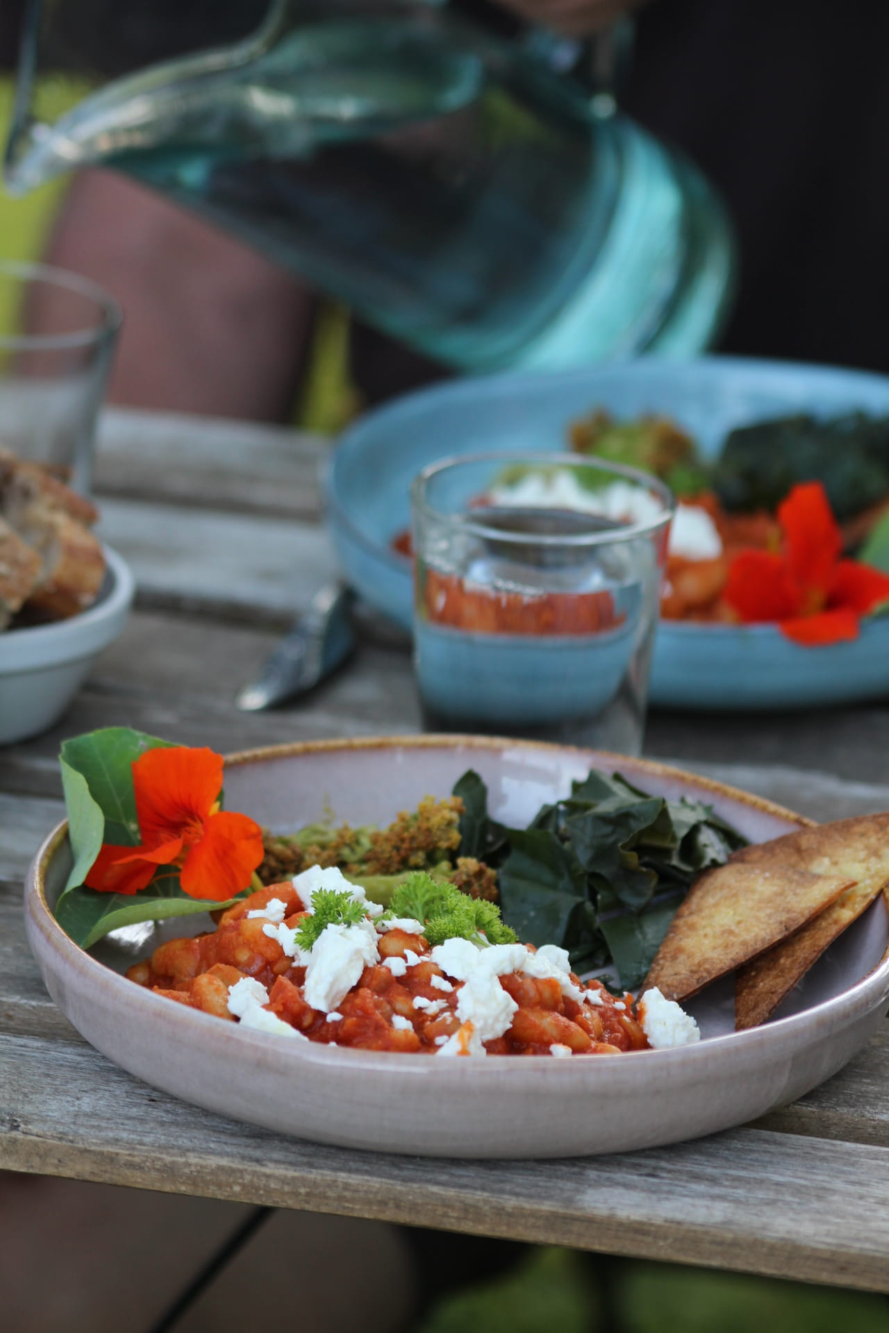 Close-up of a glass of clear liquid with a sprig of herbs, surrounded by blurred glasses on a table, creating a cozy dining atmosphere.