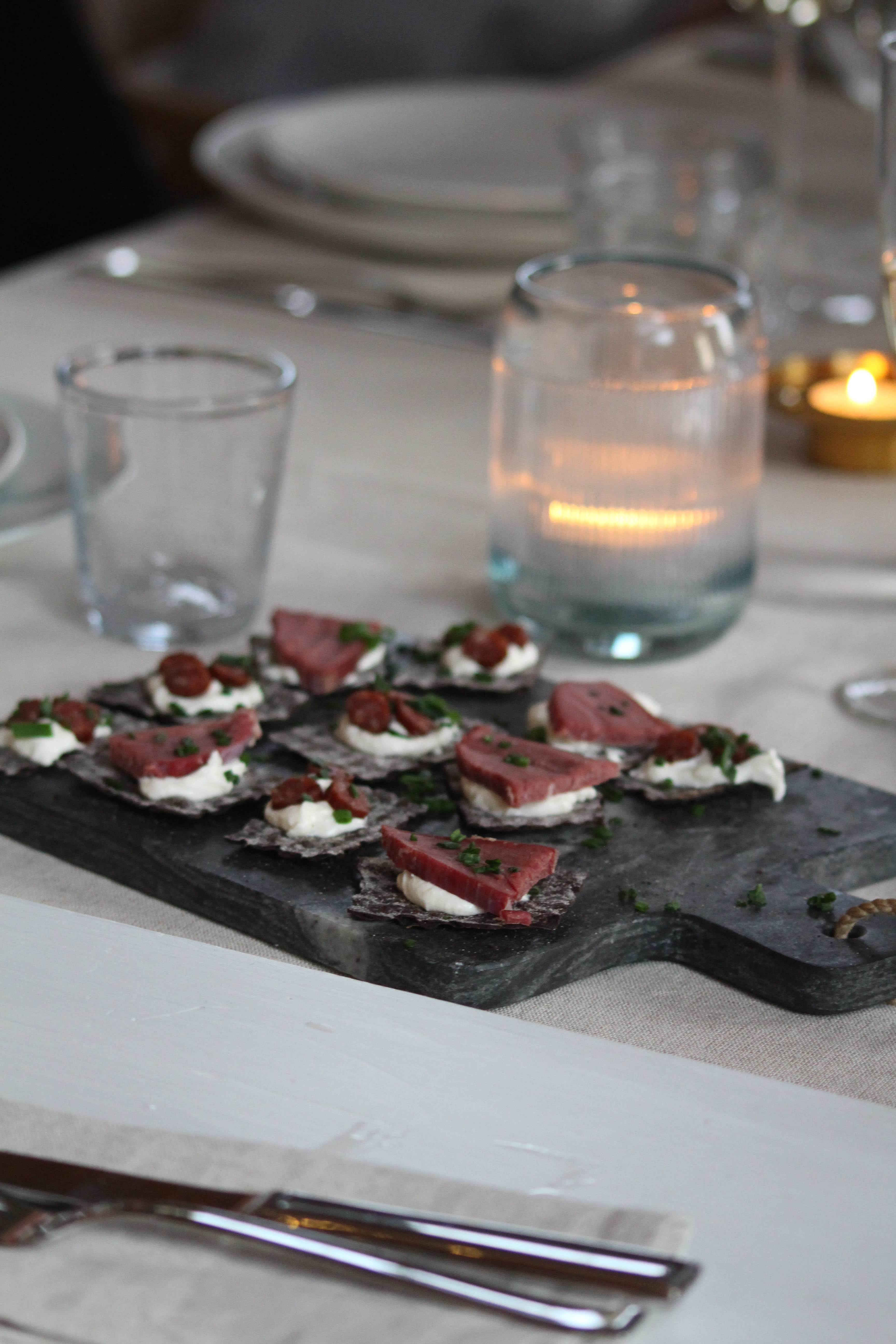 Close-up of a glass of clear liquid with a sprig of herbs, surrounded by blurred glasses on a table, creating a cozy dining atmosphere.