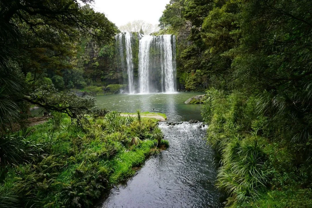 A stunning waterfall cascades into a serene pond, surrounded by lush greenery and tall trees, creating a tranquil natural scene.