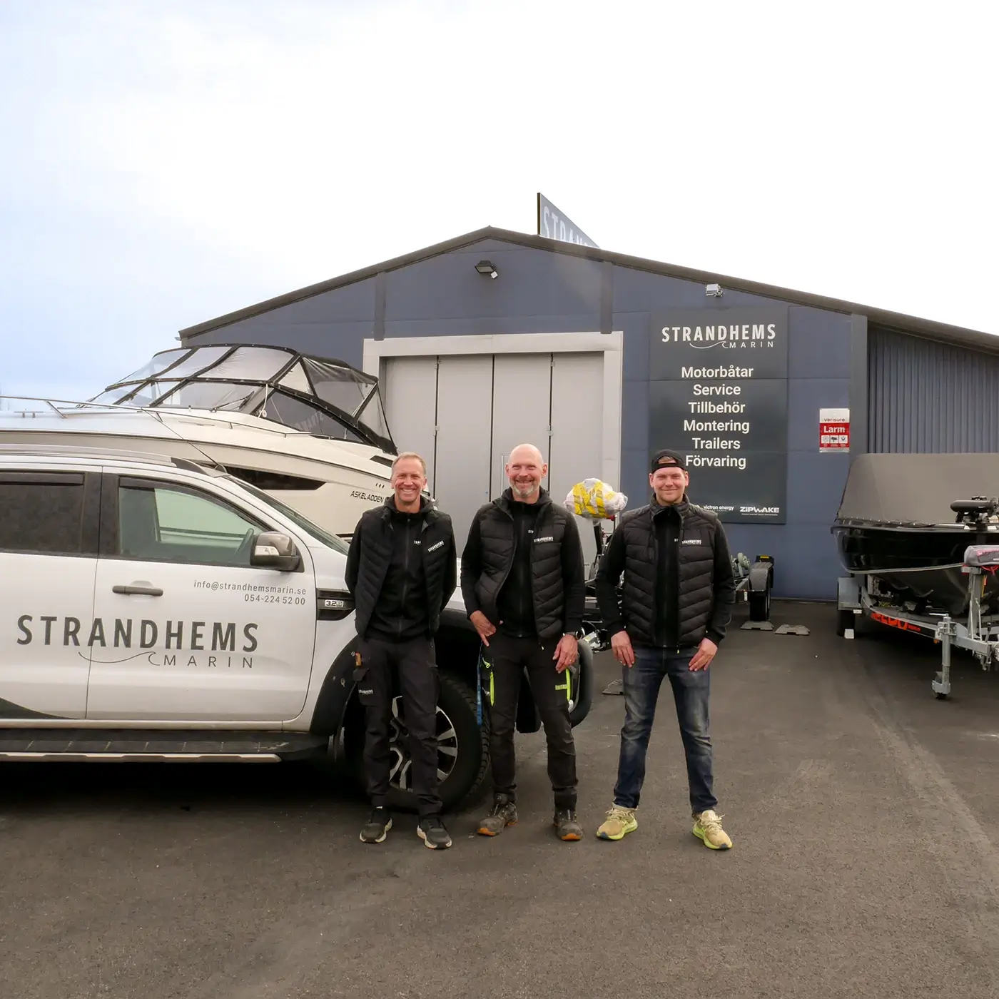 Tre män i matchande svarta västar står framför en Strandhems Marin-skylten och en vit pickup med företagets logotyp, med båtar i bakgrunden.