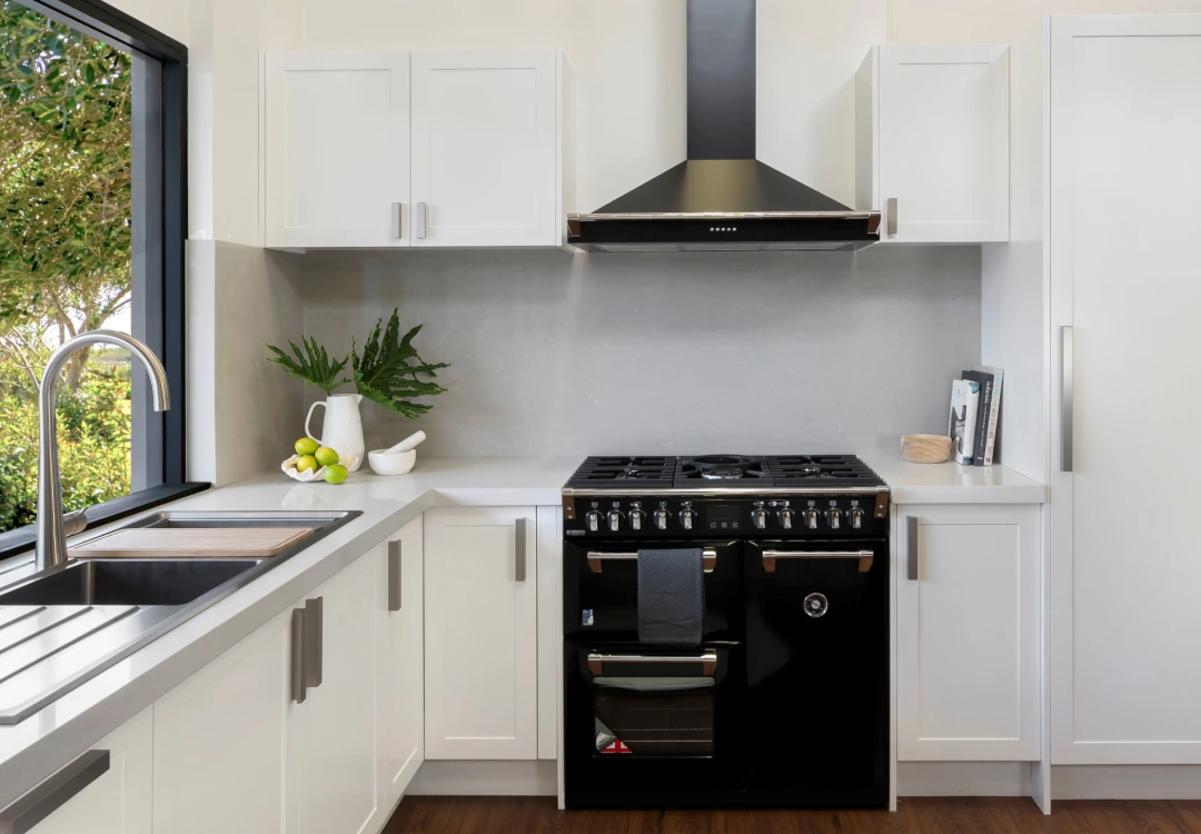 Modern kitchen with white cabinets, black stove with oven, stainless steel hood, and a window above the sink showing greenery outside.
