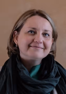 Portrait of a smiling woman with short light brown hair, wearing a black scarf and green top against a plain brown background.
