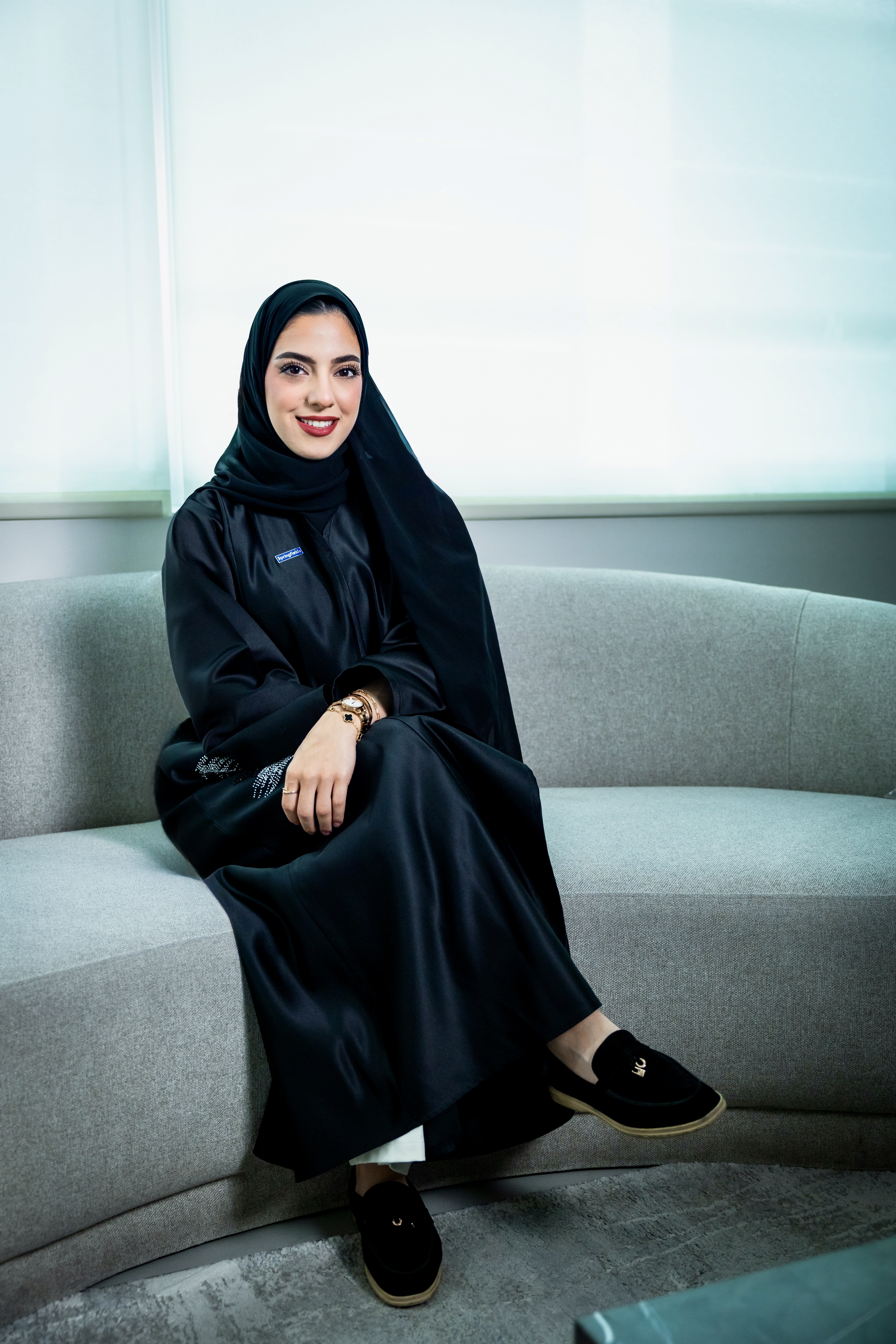 Woman in black abaya and hijab sitting on a curved gray couch smiling at the camera.