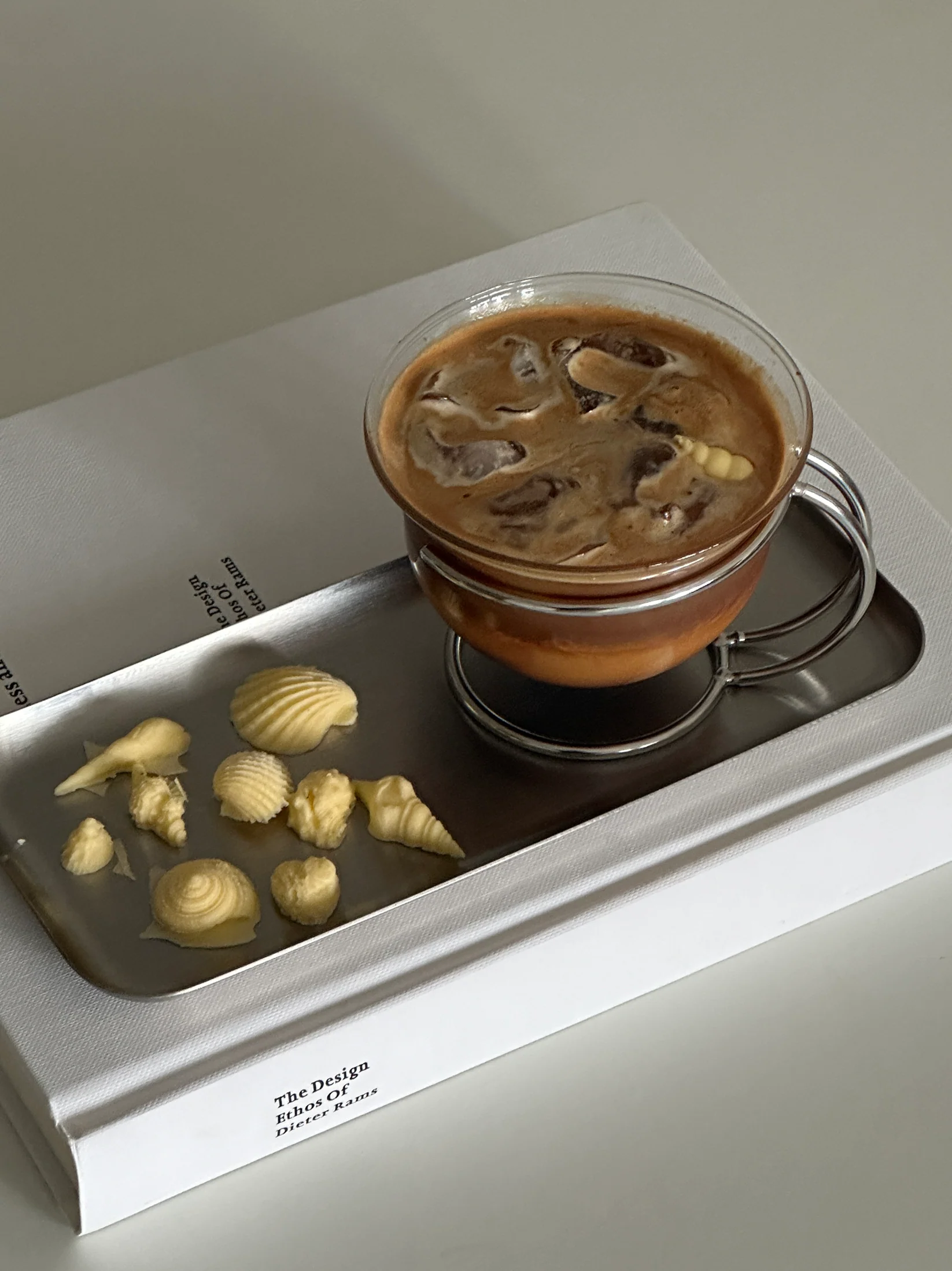 Glass cup of iced coffee on a metal tray with yellow seashell-shaped butter pieces on a white book titled 'The Design Ethos of Dieter Rams'.