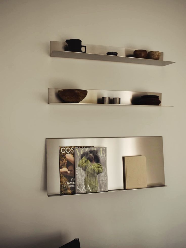 Three metal wall shelves holding cups, bowls, and magazines against a beige wall.