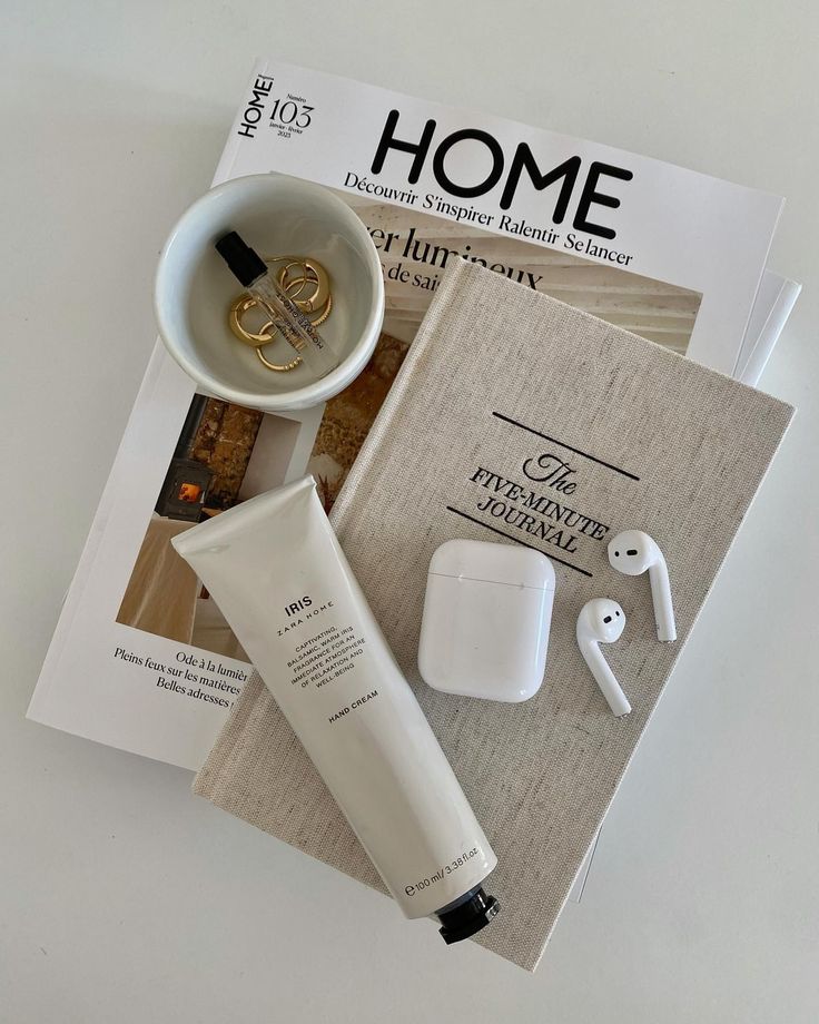 Flat lay of a beige journal, white AirPods with case, Zara Home Iris hand cream, gold rings in a small white bowl, and a HOME magazine on a white surface.