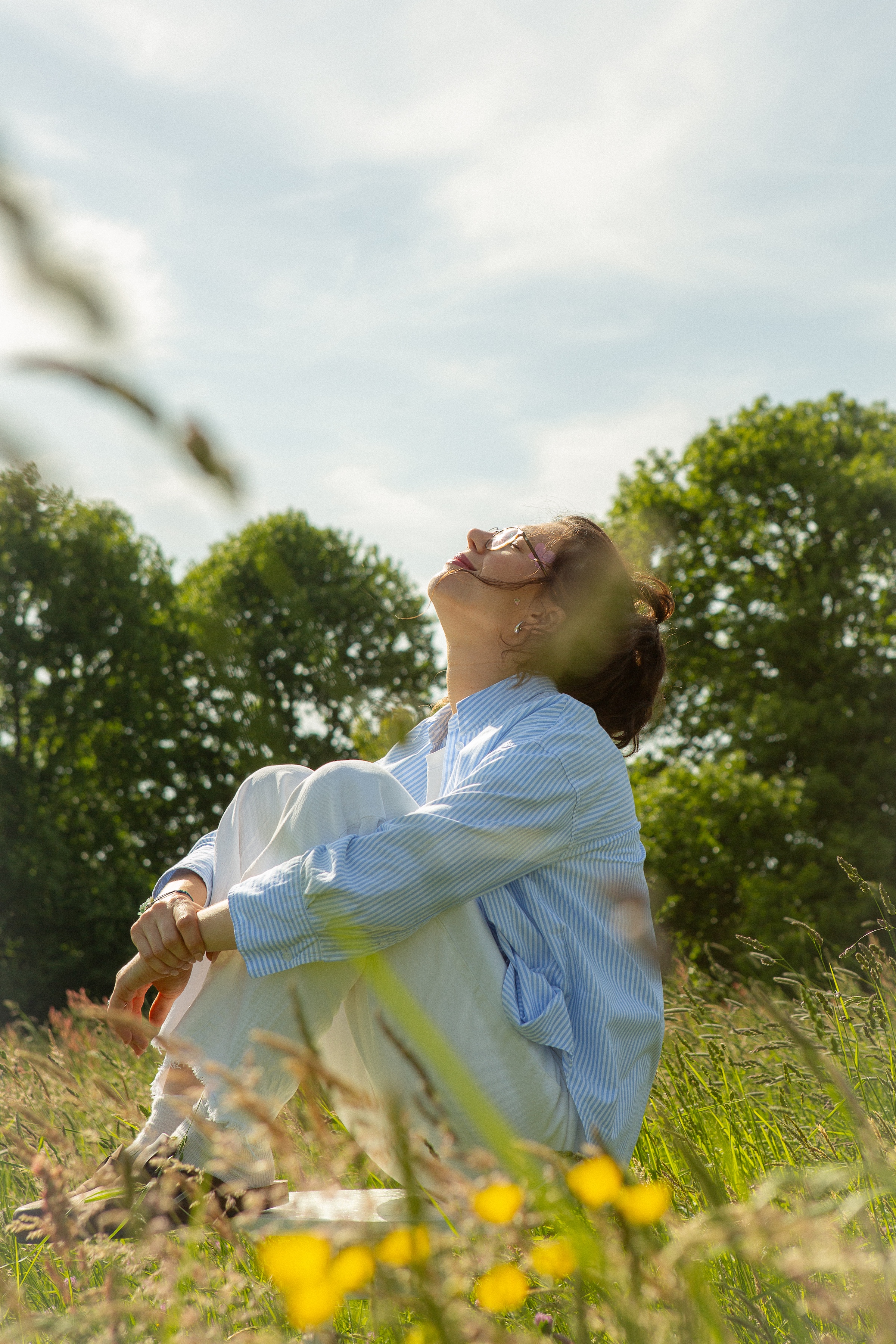 Woman sitting cross-legged in a grassy field, looking up with eyes closed, wearing glasses and a blue striped shirt.