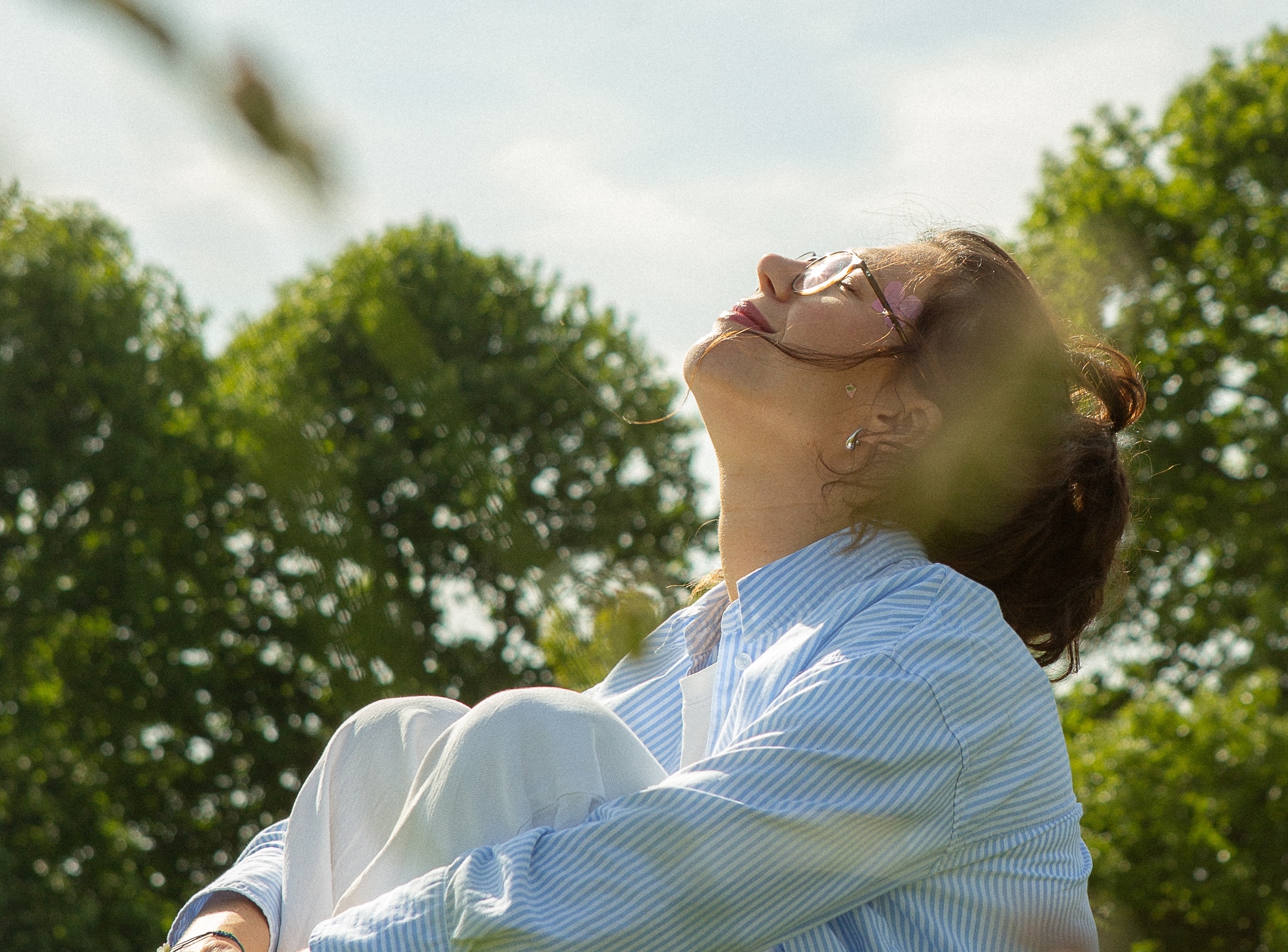 Woman wearing glasses and a blue-striped shirt sitting outdoors with eyes closed, enjoying sunlight with trees in the background.