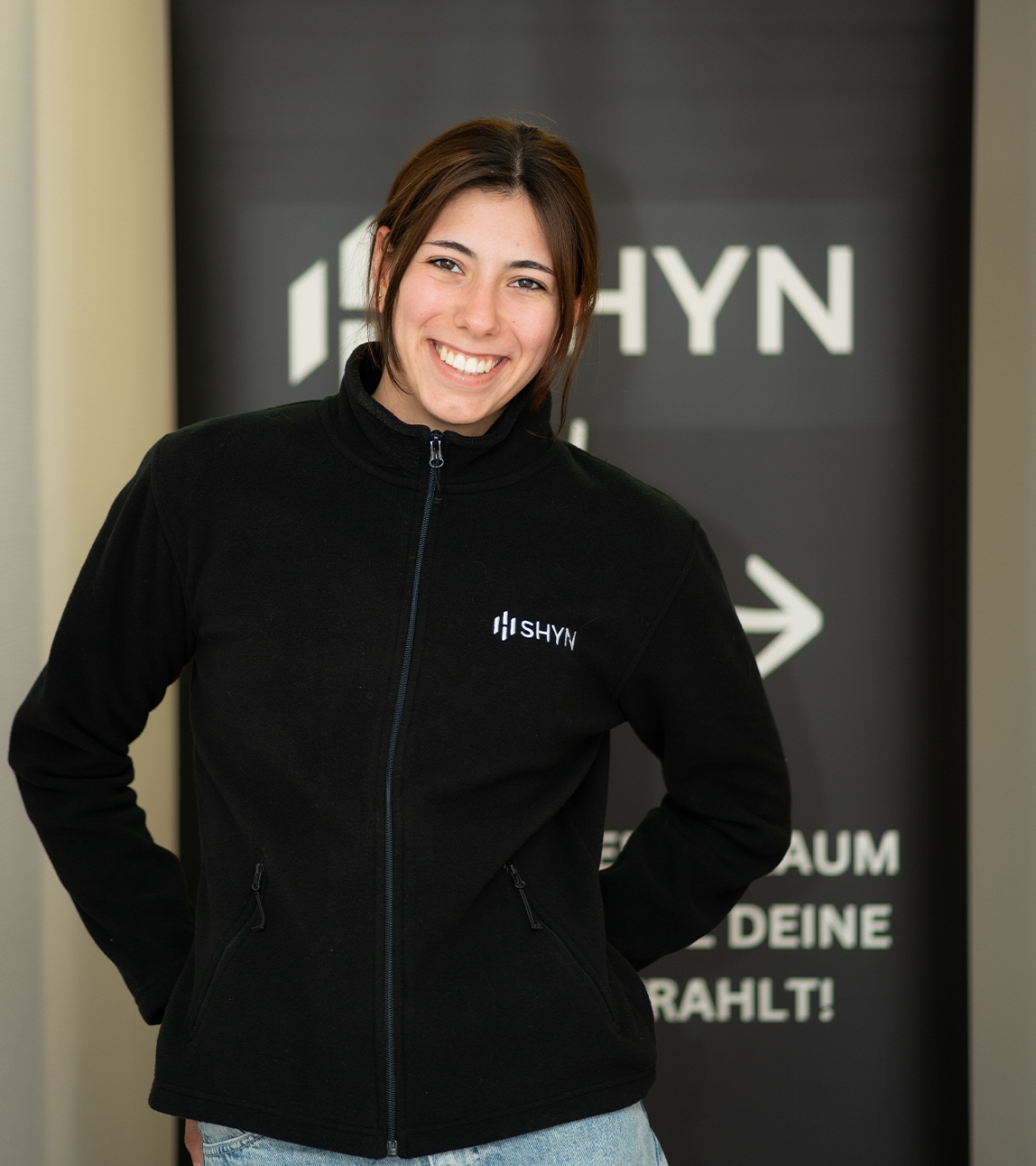 Smiling young woman wearing a black SHYN zip-up jacket standing in front of a SHYN sign.