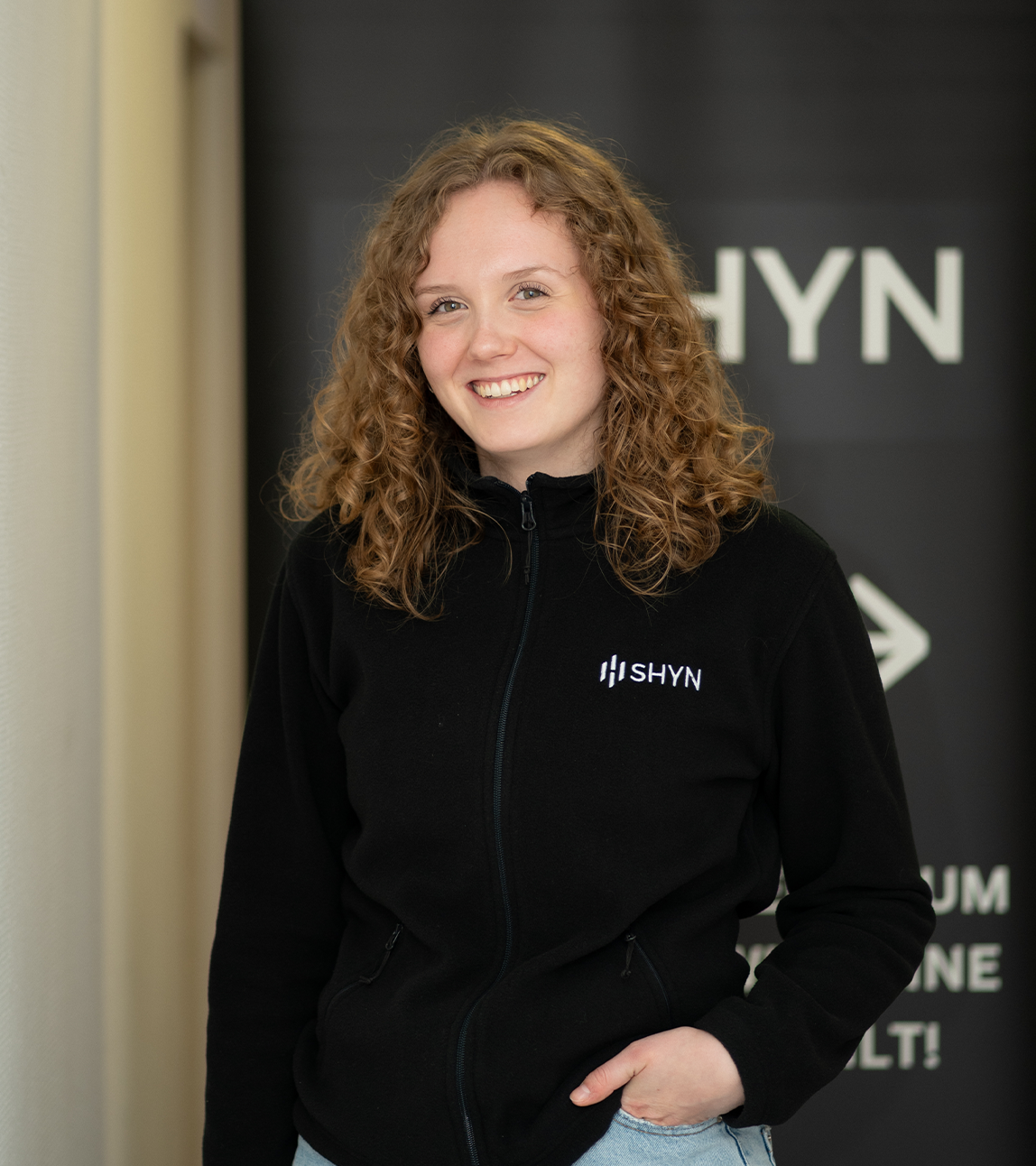 Smiling woman with curly hair wearing a black SHYN jacket standing indoors.