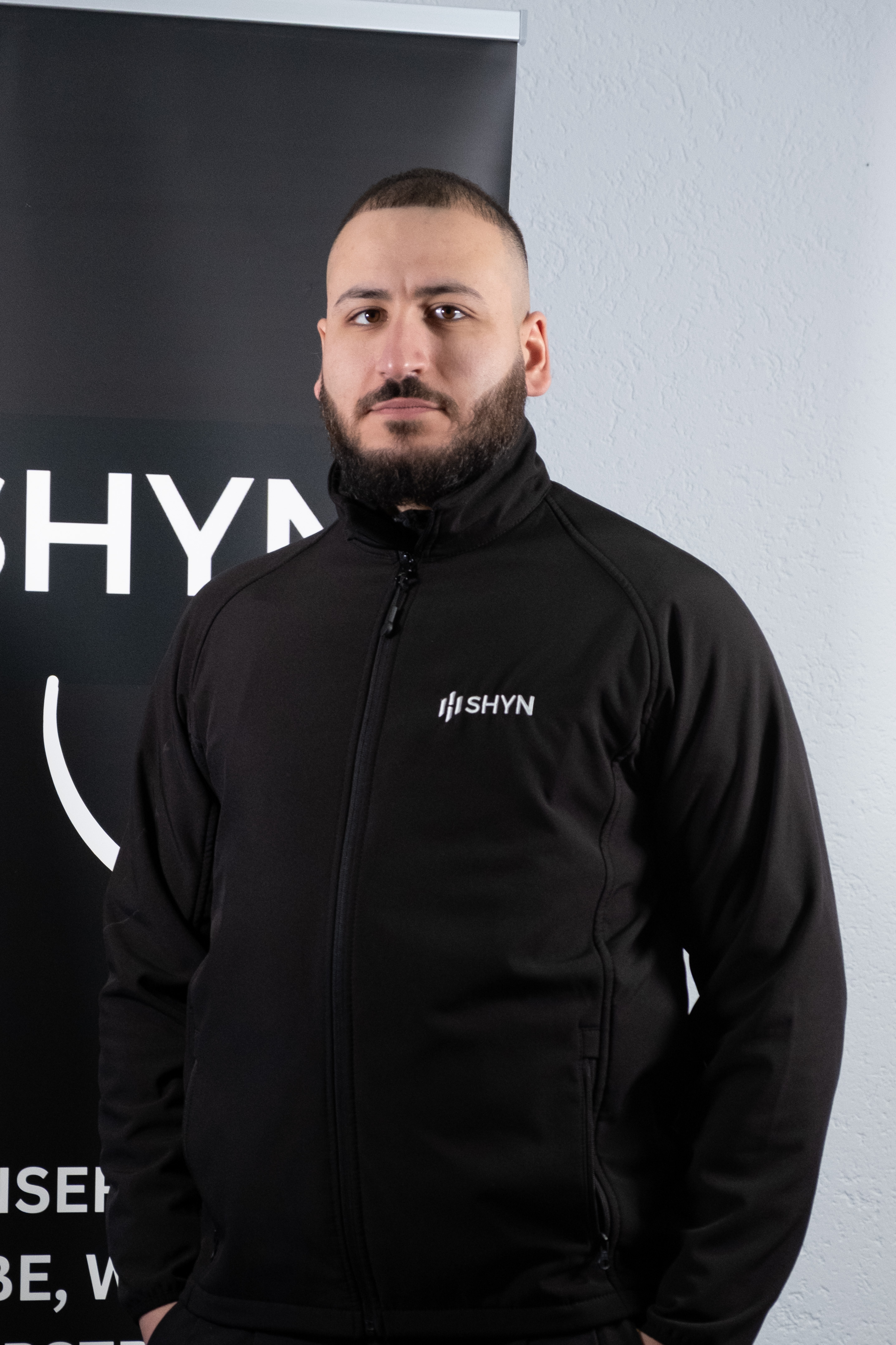 Smiling young woman wearing a black SHYN zip-up jacket standing in front of a SHYN sign.