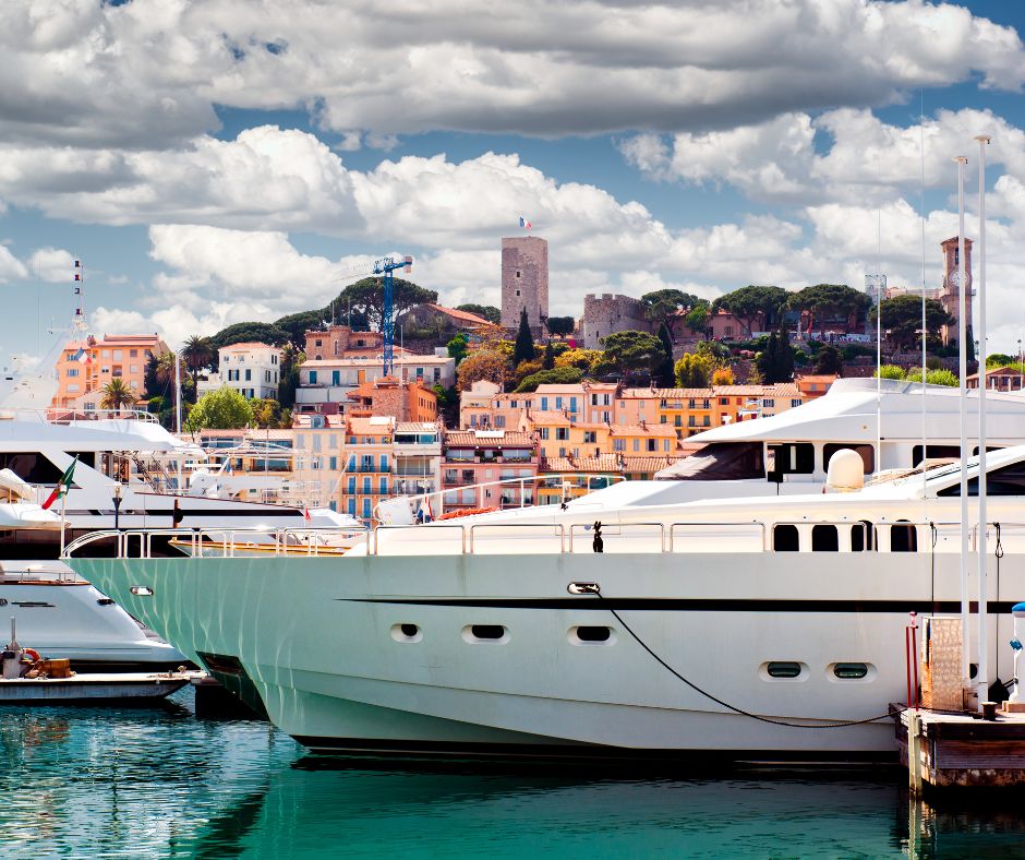 White luxury yachts docked in a marina with a coastal town and cloudy sky in the background.