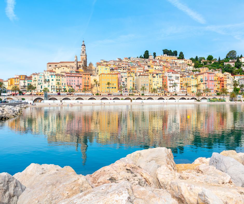 Colorful coastal town with pastel buildings reflected in calm blue water under a clear sky.