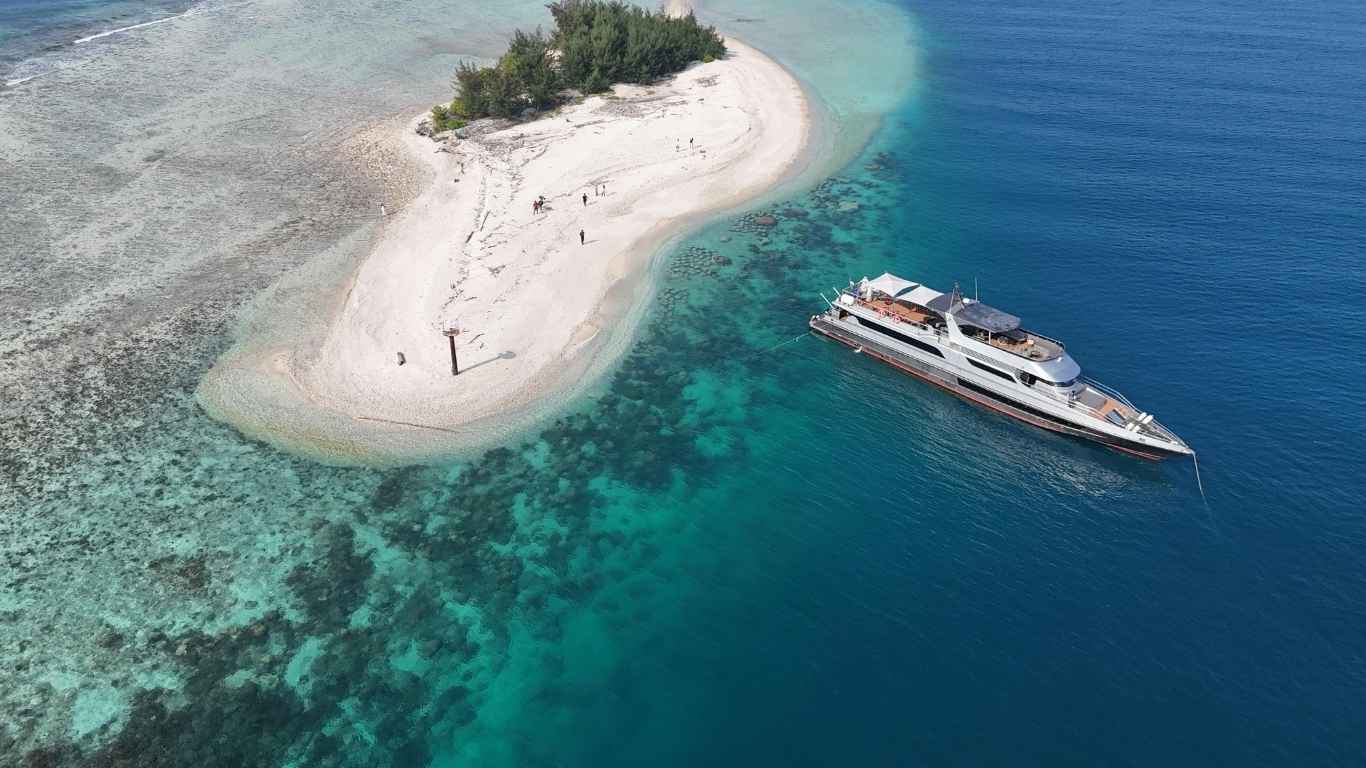 Aerial view of a small sandy island with sparse trees and people near a large anchored yacht in clear blue ocean waters.