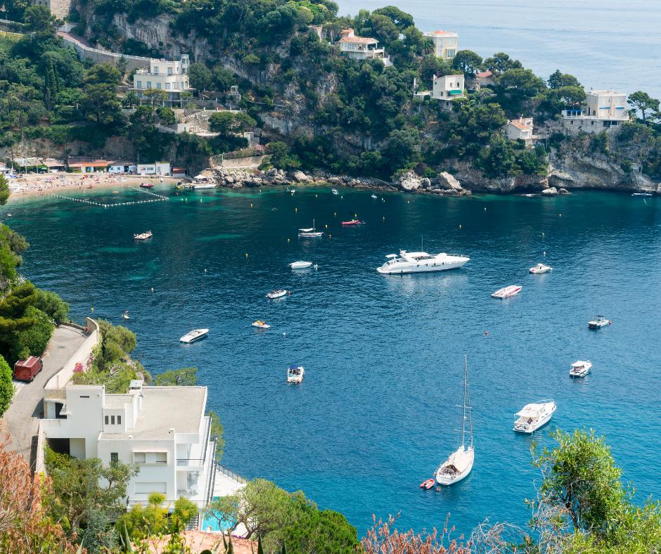 Coastal bay with multiple boats and yachts anchored in clear blue water, surrounded by green hills with houses and a small beach.