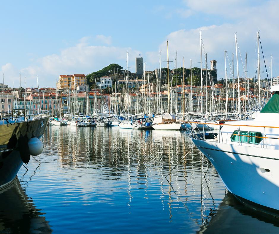 Marina with numerous sailboats docked and two large boats in the foreground, with a coastal town and castle on a hill in the background under a blue sky.