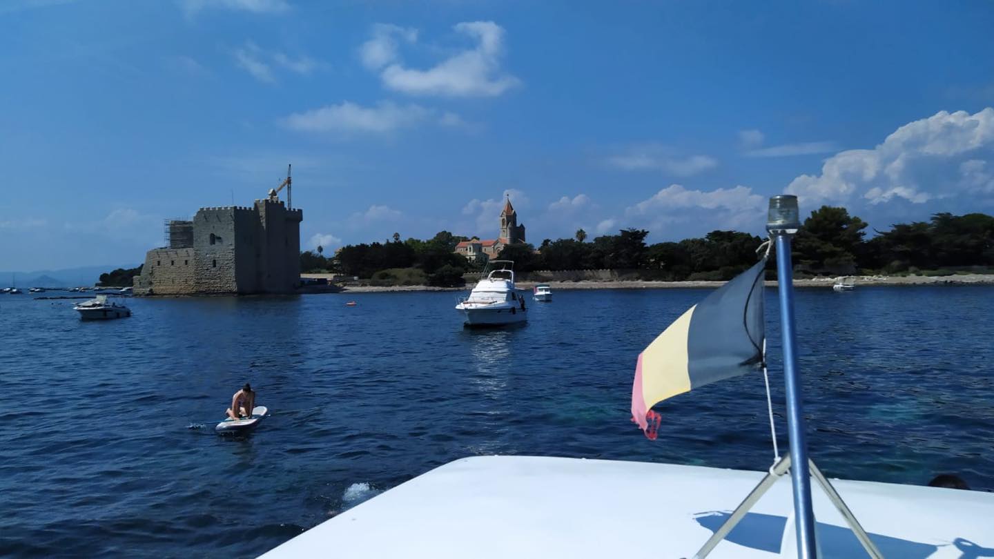 View from a boat with a tricolor flag, showing a paddleboarder on blue water and an old stone castle and church on a tree-lined coastline under a blue sky.